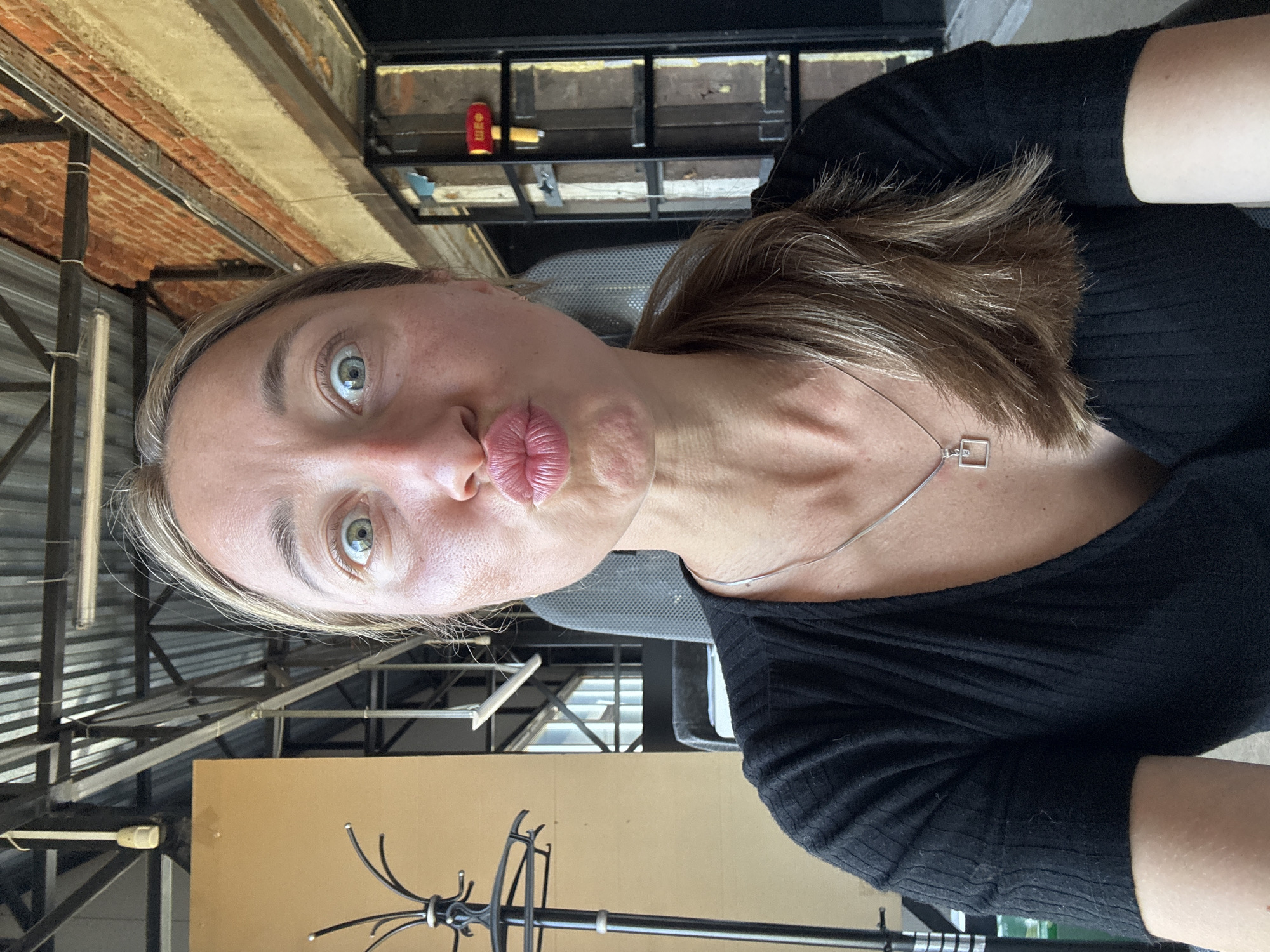 A woman with blonde hair pulled back in a ponytail, her mouth pursed in a kiss and eyes wide, poses for a selfie indoors. She is wearing a black ribbed t-shirt and a silver pendant necklace with a square charm. The background appears to be an industrial-style space with exposed beams, metal structures, and brick walls, reminiscent of a workshop or loft. A coat rack stands to the left of the woman, and what appears to be a fire axe is mounted on the wall to the right. The overall setting suggests an urban, possibly renovated industrial area within Chaikovsky, Russia.