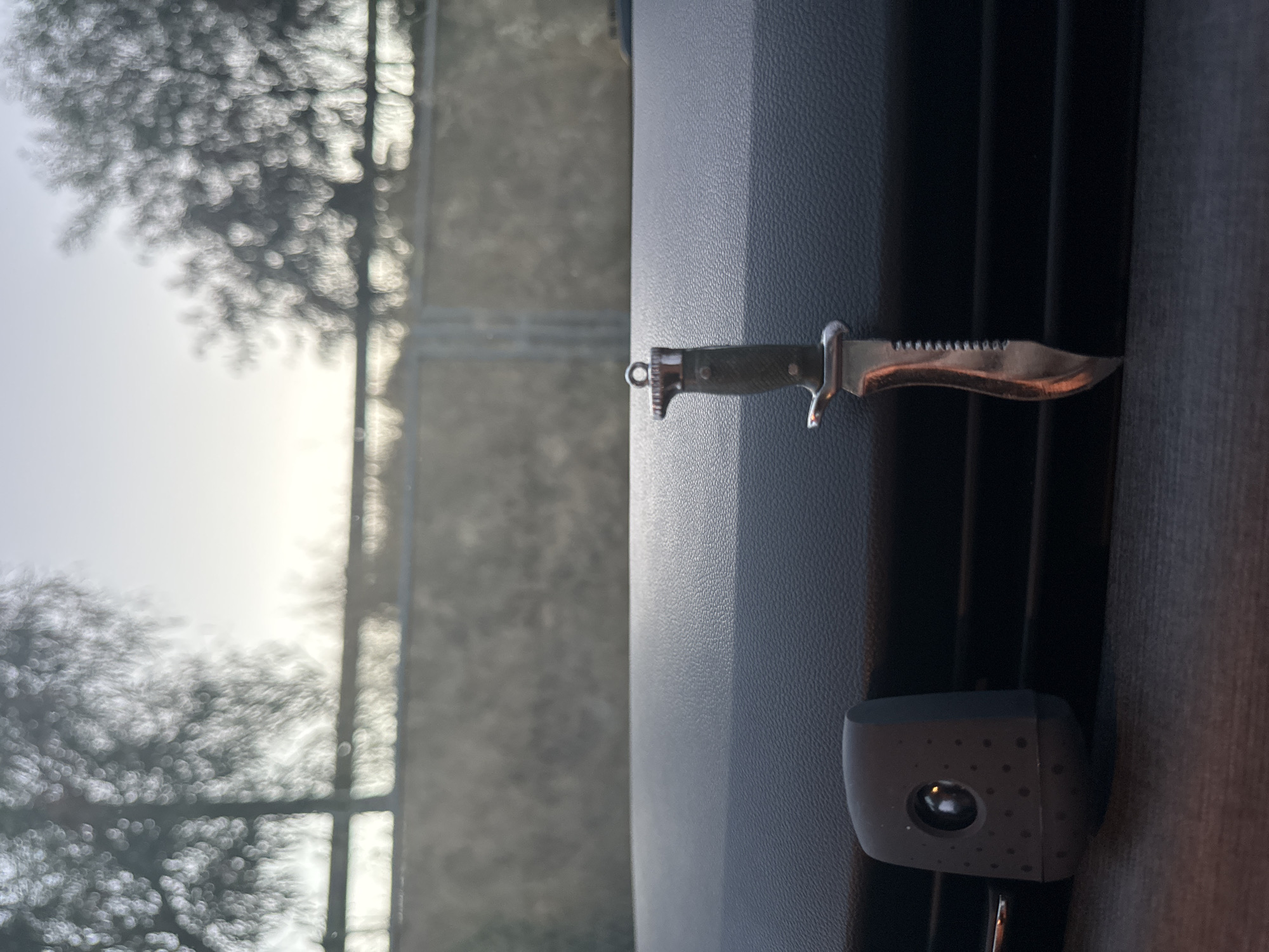 A small knife keychain hangs from the dashboard of a vehicle. The keychain is dark green with a silver blade. To the left of the keychain, a gray car air freshener is visible. The dashboard is dark gray and textured. Through the windshield, a blurred outdoor scene is visible, with trees and a fence. The lighting suggests it is either sunrise or sunset. There is no indication of the location being in the city of Murcia, Spain.