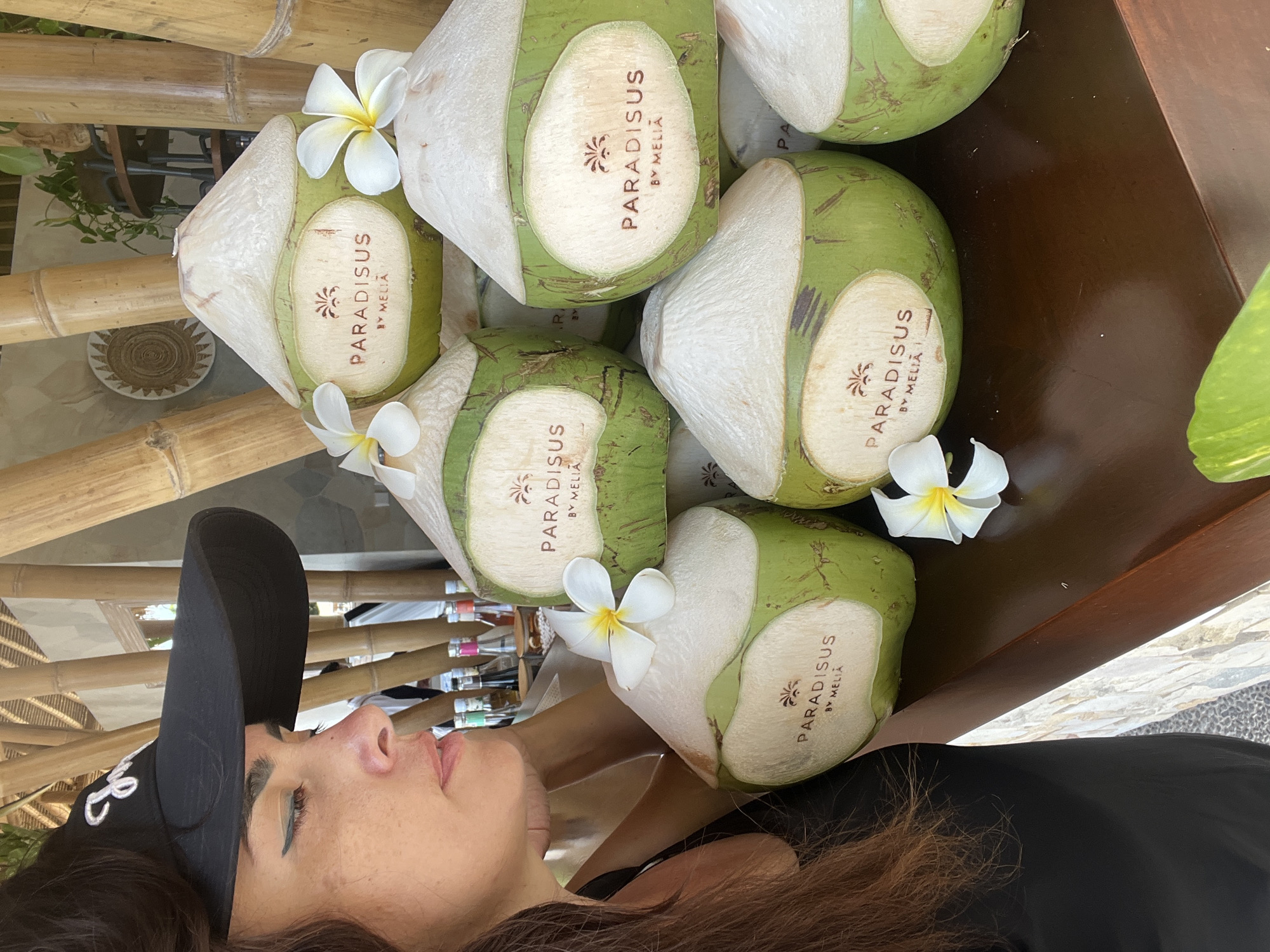 A woman wearing a black cap with white lettering is pictured in the foreground, looking to her left at a stack of green coconuts. The coconuts are branded with the logo "PARADISUS BY MELIÁ" and decorated with white plumeria flowers. Behind the coconuts, bamboo stalks are visible, suggesting an outdoor or tropical setting. The background includes a shelf with bottles, plants, and a woven circular decorative item. The overall scene conveys a resort or bar atmosphere, consistent with the Paradisus by Meliá brand, which has properties in locations like Kuta Selatan, Indonesia.
