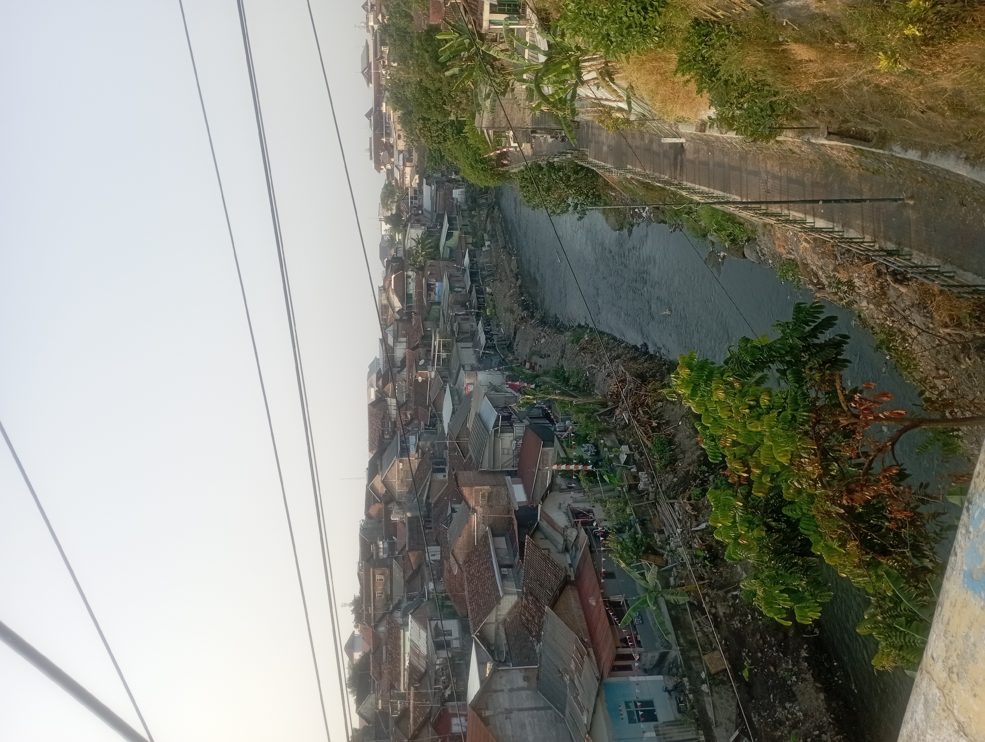 This image captures a dense urban neighborhood in Yogyakarta, Indonesia, characterized by a river flowing through it. The river, appearing murky, is flanked by a pathway on the right, lined with a metal railing and overgrown vegetation. The left bank of the river is cluttered with houses, many with red tile roofs, built close together and extending up a hillside. Some houses have visible electrical wires and antennas. Banana trees and other greenery are scattered throughout the residential area and along the riverbanks. In the distance, more densely packed buildings and trees are visible, suggesting a sprawling urban landscape. The sky is overcast, creating a soft, diffused light over the scene. A few people are visible walking on the pathway beside the river.