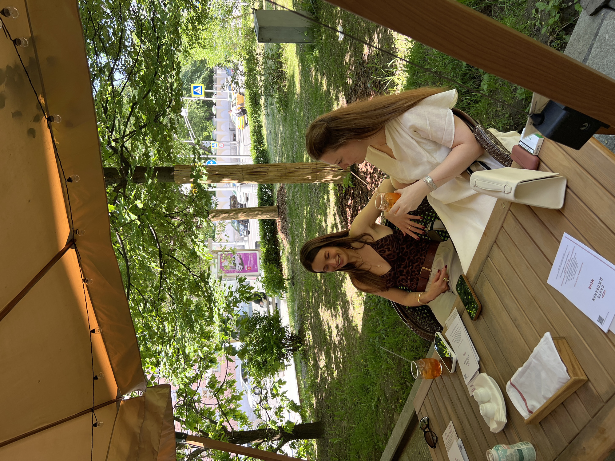 The photograph depicts two women at a table in an outdoor setting. The woman on the right is fair-skinned, with long brown hair, wearing a white blouse. She is holding a glass with an orange-colored drink and appears to be looking at the other woman. The other woman, on the left, has dark hair and is wearing a brown leopard-print top. She is smiling, with her hand resting on the table next to her smartphone.
The table is wooden and cluttered with items including menus written in Russian, napkins, sunglasses, and what appears to be a porcelain figurine. Drinks, including the one the woman on the right is holding, are also visible on the table.
The background consists of greenery, including grass and trees, suggesting they are in a park or outdoor cafe setting. Across the street are buildings and street signs. Based on the style of the buildings and signage, the location is likely in Moscow, Russia. An open umbrella or canopy is partially visible on the left side of the frame. The scene is bathed in natural light, likely during daytime.