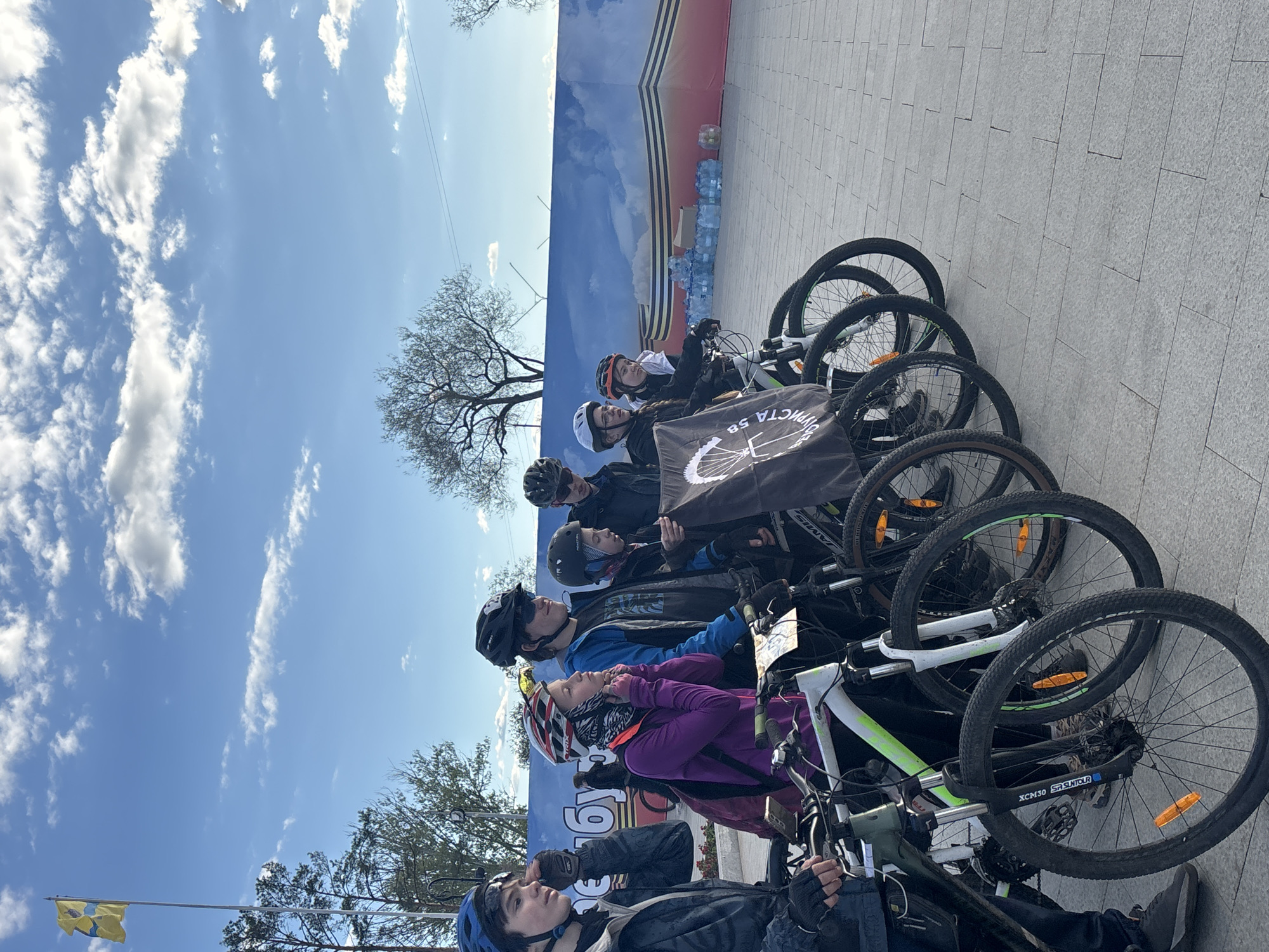 A medium, outdoor, eye-level shot captures a group of people with their bicycles standing on a grey brick pavement. The setting is identified as Orenburg, Russia. At least six people are present, all wearing helmets and casual clothing, suggesting they may be part of a cycling event or tour. Each person is standing next to their bicycle, which are closely aligned along the pavement. A flag, possibly indicating a cycling group or club, is visible near the center. In the background, a large banner or backdrop displays a sky and a city scene, potentially representing the local landscape of Orenburg. The sky above is clear with scattered clouds, indicating a sunny day. The overall composition suggests a staged or organized gathering of cyclists in an urban environment.