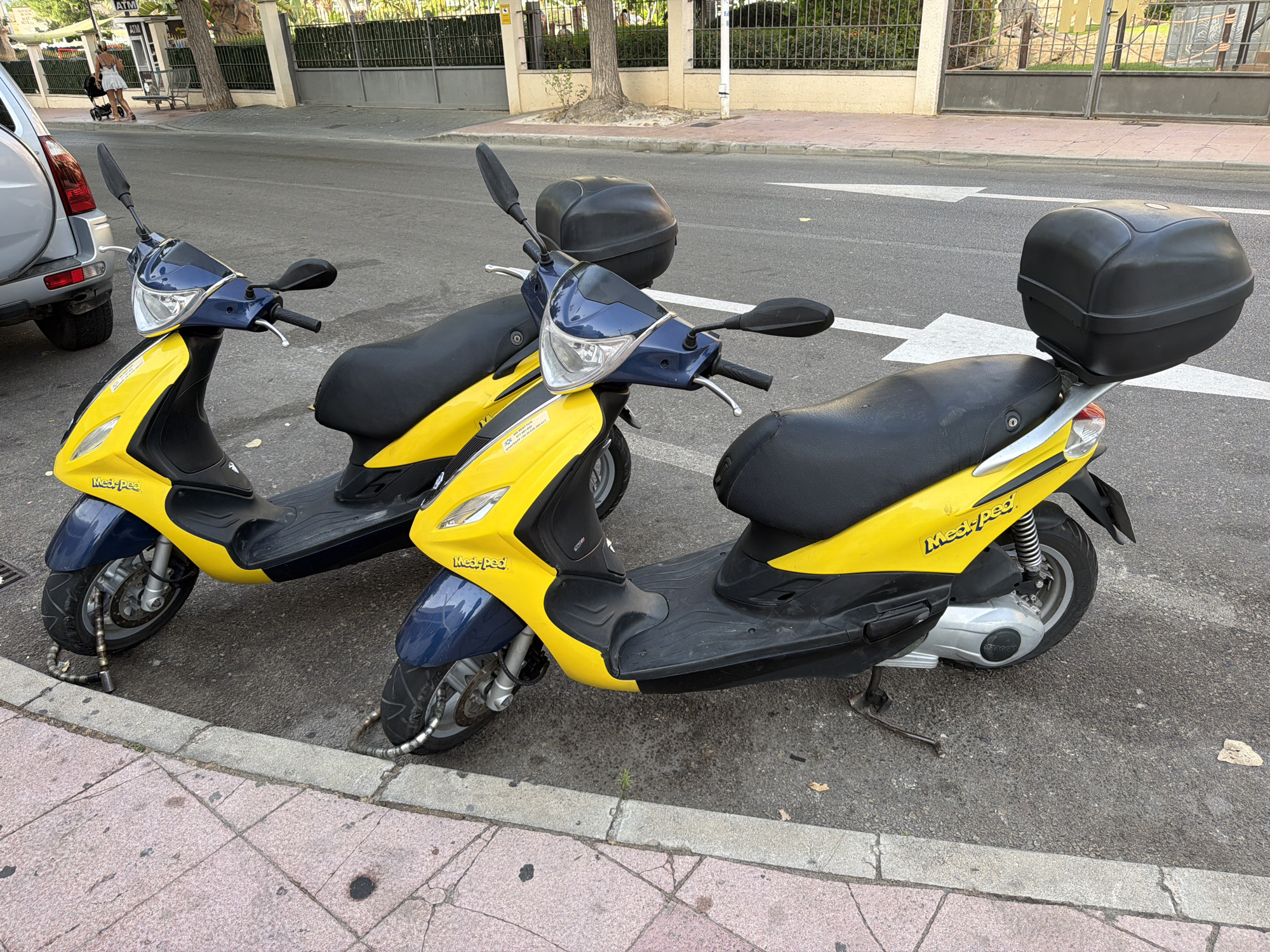 Two yellow and blue scooters are parked on the side of a street in Magaluf, Spain. The scooters are chained to the curb with thick metal chains. The scooter on the left is closer to the camera and is partially obscured by a silver SUV. The scooter on the right is behind the first scooter and has a black storage box on the back. The street is paved, and there is a sidewalk with pink tiles. In the background, there are trees, a fence, and buildings. A woman with a stroller is walking on the sidewalk in the distance.