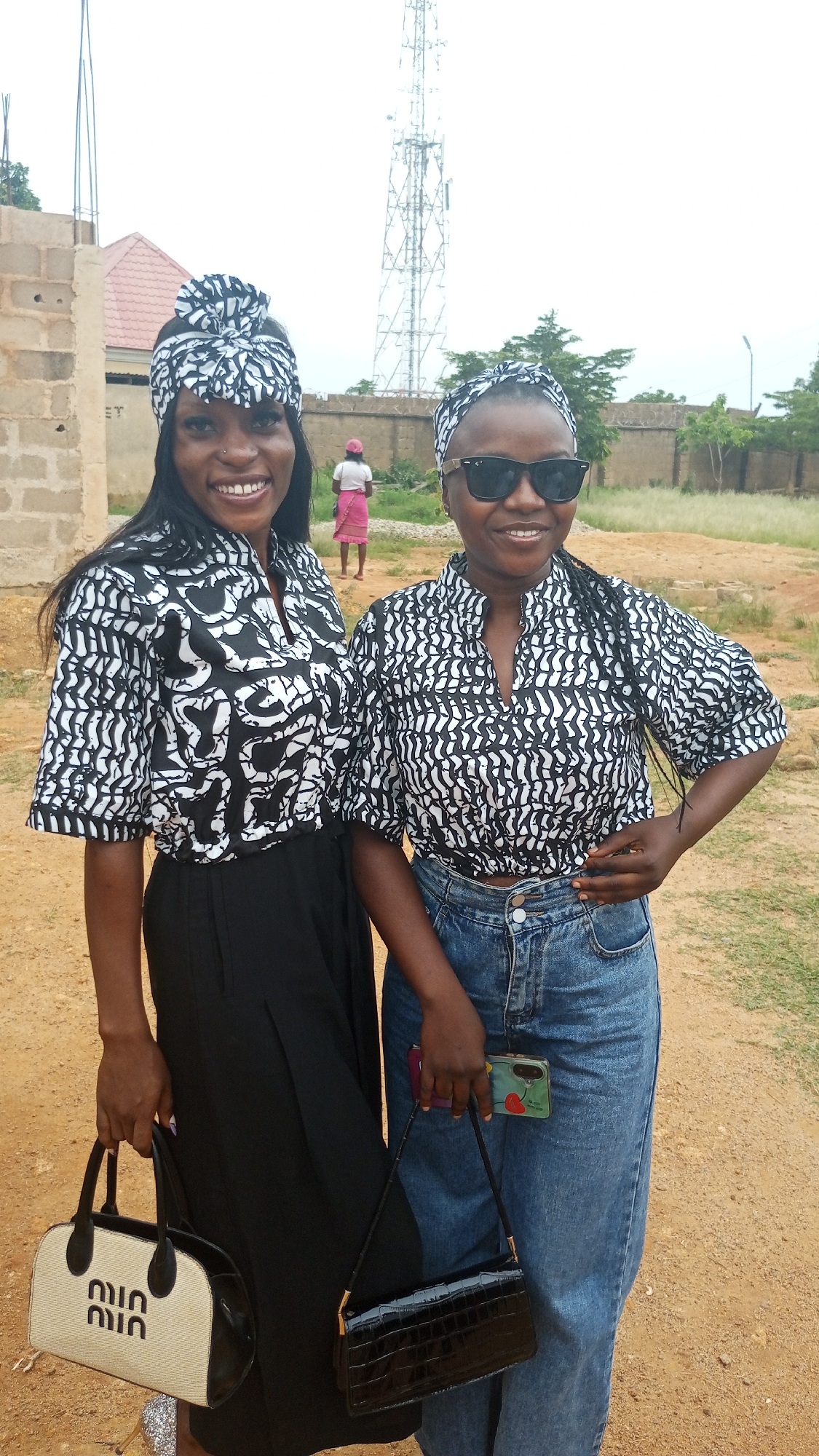 Two women stand on a dirt path in Barikin Sabon Gidan Kanat, Nigeria. They are both wearing matching black and white patterned shirts and head wraps. The woman on the left wears black pants and holds a beige and black purse. The woman on the right wears blue jeans, sunglasses, and holds a black purse and a phone. In the background, there is a partially constructed brick building, a telecommunications tower, a fence, and a few trees. A woman in a pink outfit is walking in the distance.