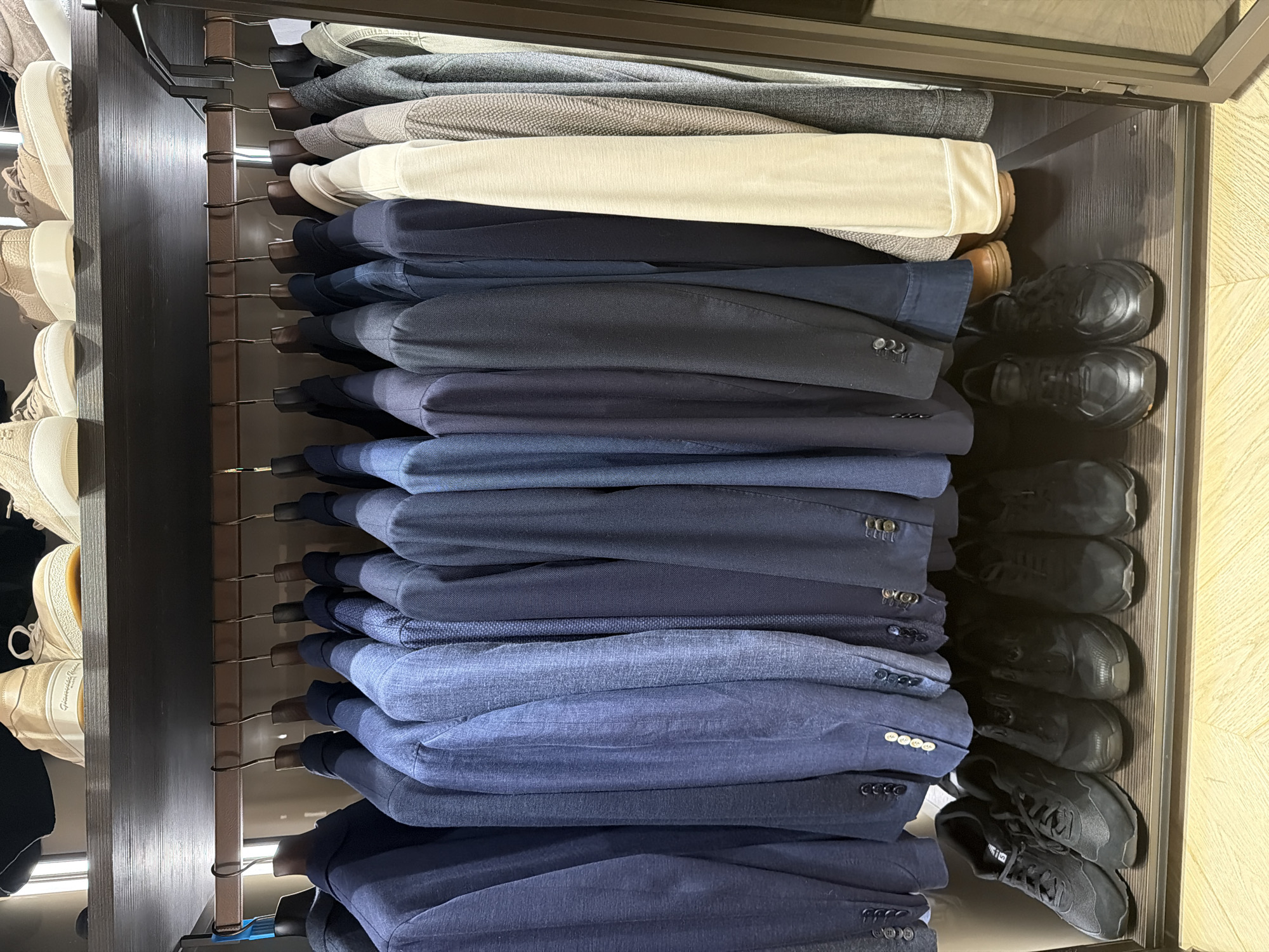 The image shows a closet with men's clothing and shoes. On the top shelf, there are several pairs of light-colored sneakers. Below, a row of blazers in various shades of blue and gray are hanging on hangers. The blazers appear to be made of different textures. On the bottom shelf, there are several pairs of black athletic shoes and one pair of brown leather shoes. The closet has dark wood shelving and is enclosed by a glass door on the right side. The environment is indoors, likely a home or retail setting. There are no people visible in the image. This scene is not associated with a specific location in Moscow, Russia.