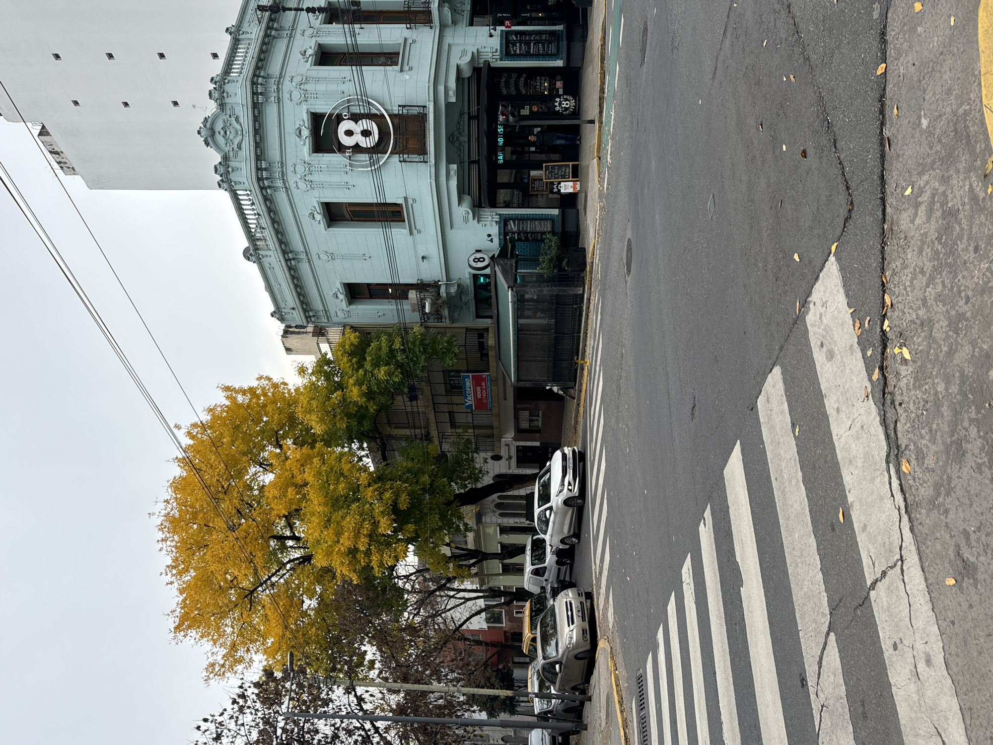 The image shows a street scene in Buenos Aires, Argentina, focused on the corner of a light blue, ornate building with the number "8" prominently displayed. This building is the "El 8 Bar Rotise", known for rotisserie-style food, as indicated by signage on the awning and sidewalk. A crosswalk is in the foreground, and parked cars line the street on the right side of the frame. A tree with yellow foliage stands to the left of the building, while other trees with bare branches are further back. The sky is overcast, and a tall, white building is visible behind the El 8 building. The street appears relatively quiet with no pedestrians visible at the crosswalk.