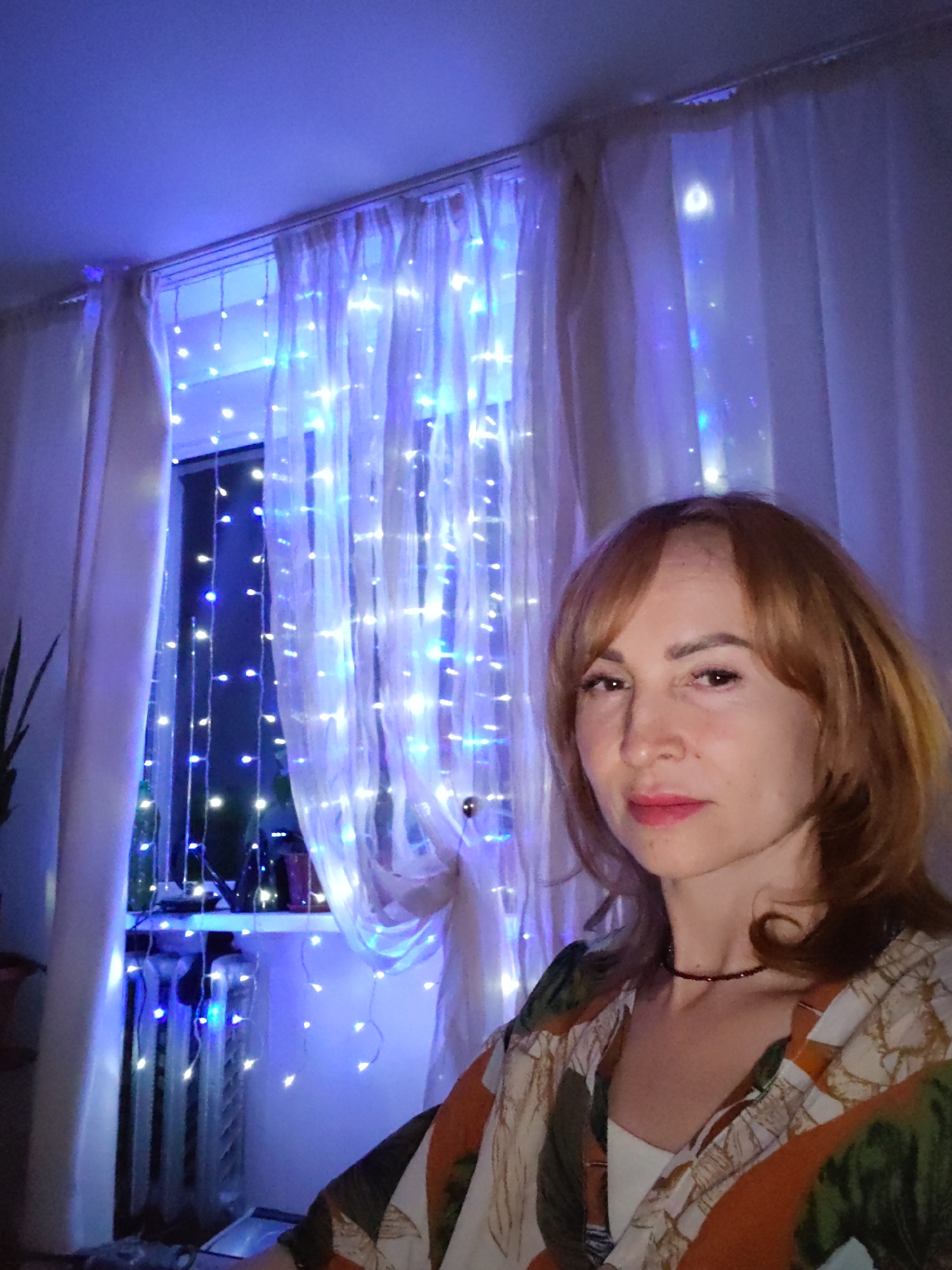 A woman with light reddish-brown hair and a floral patterned shirt is positioned in front of a window adorned with string lights and sheer curtains. The scene is bathed in a blue light emanating from the decorative lights. The window appears to be in a room in a building, likely an apartment, based on the presence of a radiator under the window and personal items on the windowsill. The woman is wearing a dark, thin necklace. The city of Chaikovsky, Russia, is mentioned as the location.