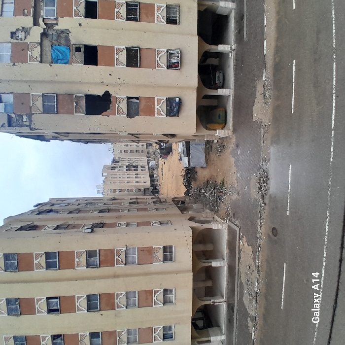 This image depicts a damaged urban residential street in Khan Yunis, Palestinian Territory, under an overcast sky. The foreground features a dark asphalt road with two parallel white dashed lines. Debris, including small rocks and dirt, is scattered on the road and along its edges.
Two multi-story apartment buildings, light beige with reddish-brown decorative window panels, flank a rubble-strewn open area. The building on the right exhibits severe damage, with multiple large sections of its facade blown out, revealing dark interiors. Numerous windows are shattered, missing, or boarded with dark material, and a blue tarp hangs from an upper-story window. Pockmarks consistent with projectile impacts are visible across its exterior. At its ground level, supported by arches, a yellow vehicle and a white sedan are parked.
The building on the left shows less extensive but still evident damage, with several windows broken or missing. Its ground level also features arches, appearing empty.
Between these two buildings, the ground is composed of dirt and significant piles of concrete, twisted metal, and general construction debris. A small, grey or white temporary structure, resembling a tent or shelter, is situated within this debris field. In the midground and background, additional multi-story residential buildings, similar in architectural style, are visible, some also appearing damaged. No individuals or overt actions are discernible in the scene.