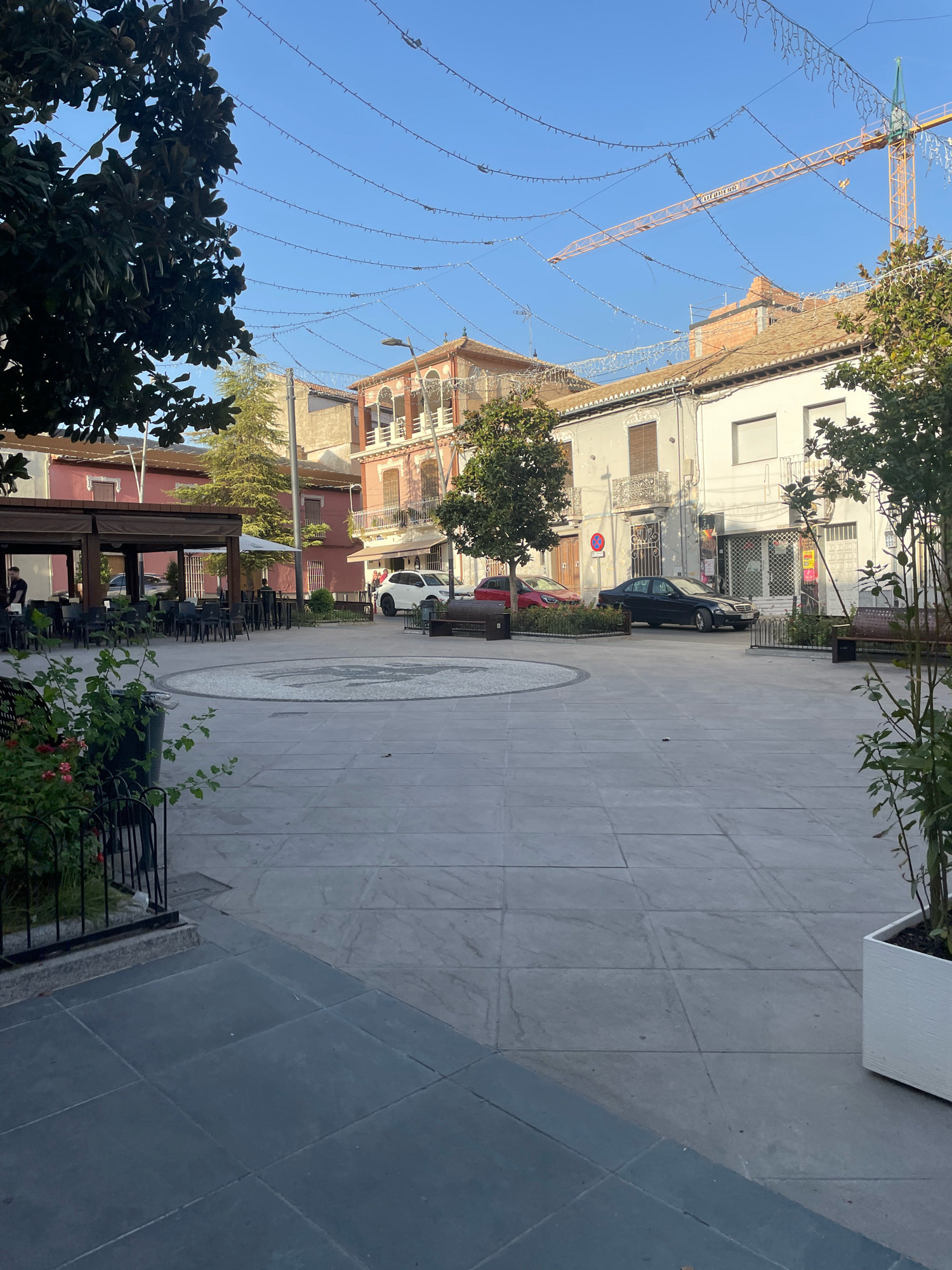 The image depicts a sunlit urban square in Atarfe, Spain, featuring a large paved area with distinct zones. In the foreground, dark gray large square tiles form a pathway, transitioning to lighter gray rectangular tiles that dominate the central part of the square. A circular decorative element with a mosaic pattern is situated in the middle of the paved area.
To the left, an outdoor seating area of a cafe or restaurant is visible, with numerous dark chairs and tables arranged under a covered structure. A few individuals are present in this area, with one person standing near the counter. Green foliage and flowering plants are also present in this section.
Surrounding the square are buildings of varying architectural styles and colors. On the left, a building with a reddish-pink facade features balconies. In the center background, a taller building with intricate details and balconies, painted in shades of ochre and white, stands out. To the right, a row of white and beige buildings with shutters and balconies line the square.
Several vehicles are parked in the square, including a white SUV, a black sedan, and a red car. A construction crane, painted yellow and orange, is visible in the background, extending above the buildings. The sky is clear and blue, with strings of small lights strung across the sky, suggesting festive decorations. Benches are placed along the edges of the square.