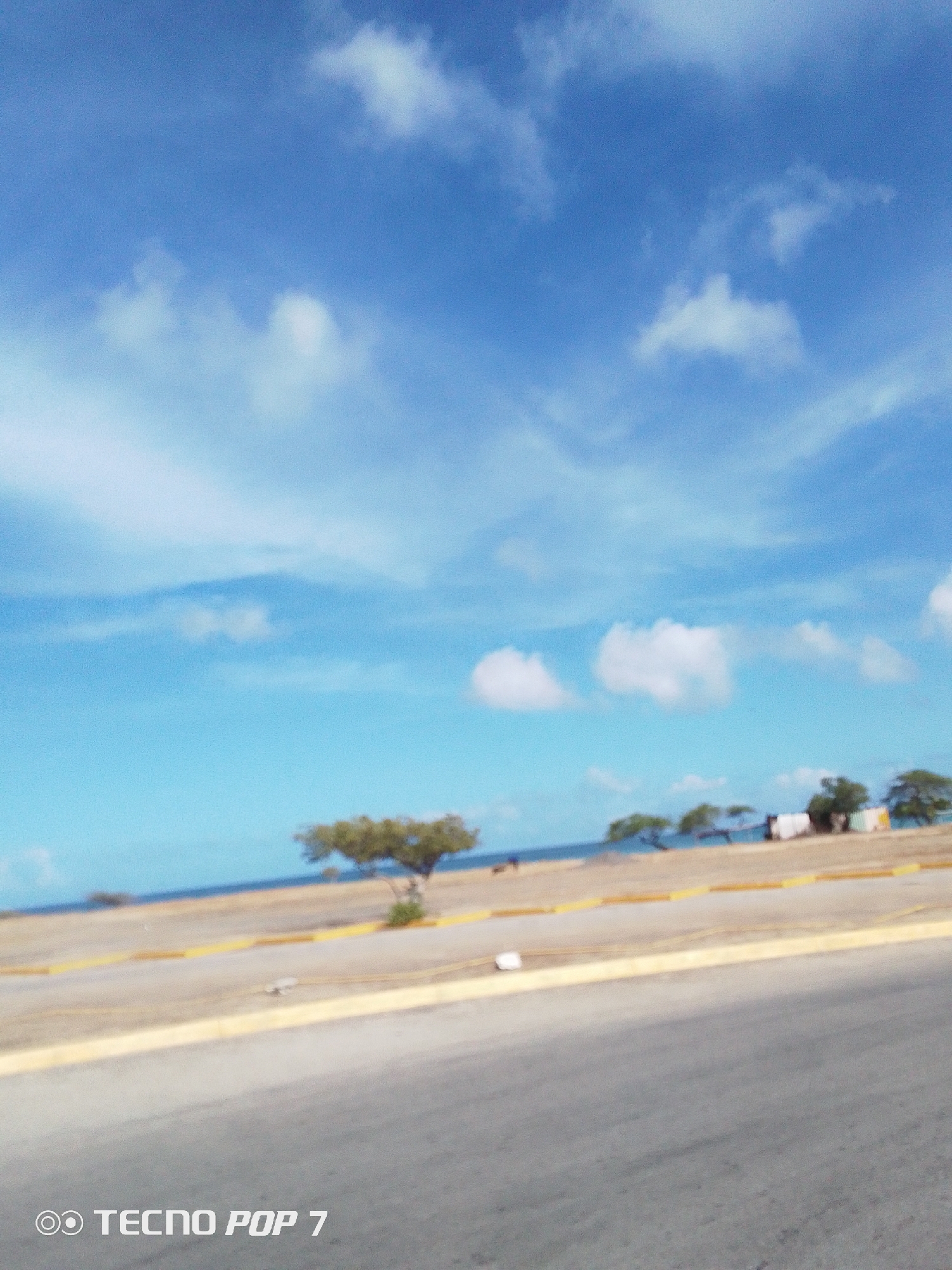 The image shows a view near the coast of Pedro González, Venezuela. A road with a yellow curb runs along the foreground. In the background, there is a sandy area that leads to the sea. Several trees are scattered across this area. Some buildings are also visible further back. The sky is mostly blue with scattered white clouds. The image is somewhat blurry. The camera is a TECNO POP 7.