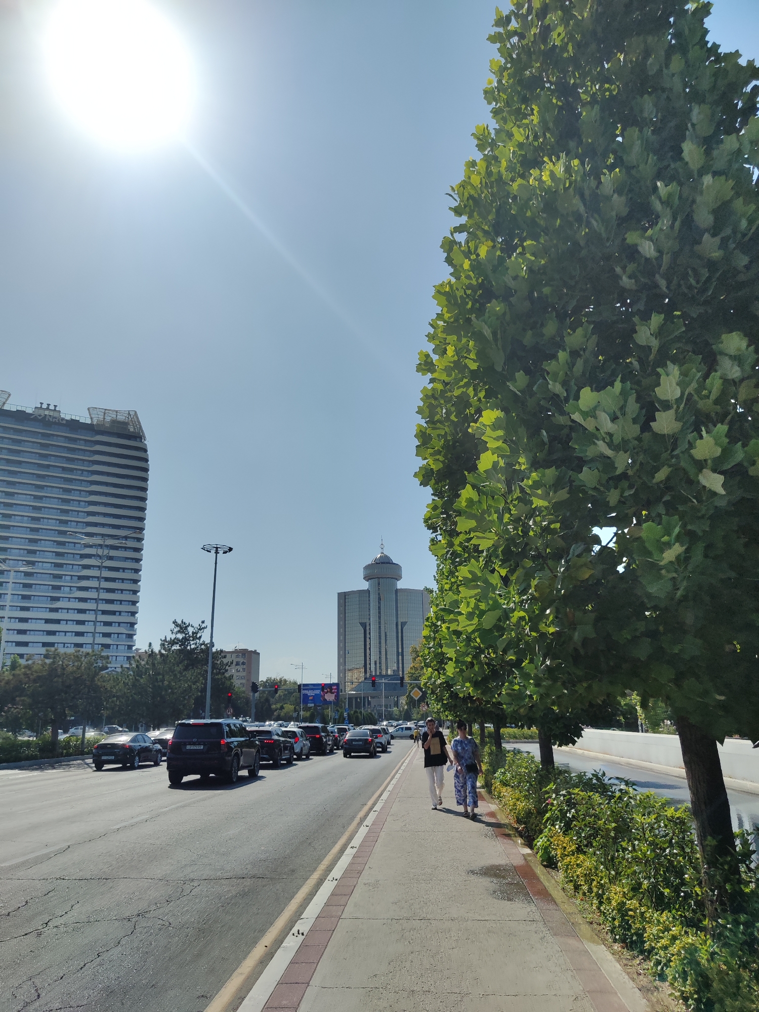 In Tashkent, Uzbekistan, this image captures a sunny day on a city street. The foreground features a sidewalk with two women walking. One woman wears a dark top and white pants, while the other is dressed in a patterned blue outfit. Lush green trees and bushes line the sidewalk. Across the street, a multi-lane road carries moderate traffic, with cars and SUVs visible. In the background, modern high-rise buildings dominate the skyline, including a tall, striped building on the left and a glass-faced building with a distinctive dome on the right. The sky is clear and bright, with the sun strongly illuminating the scene from the upper left corner.