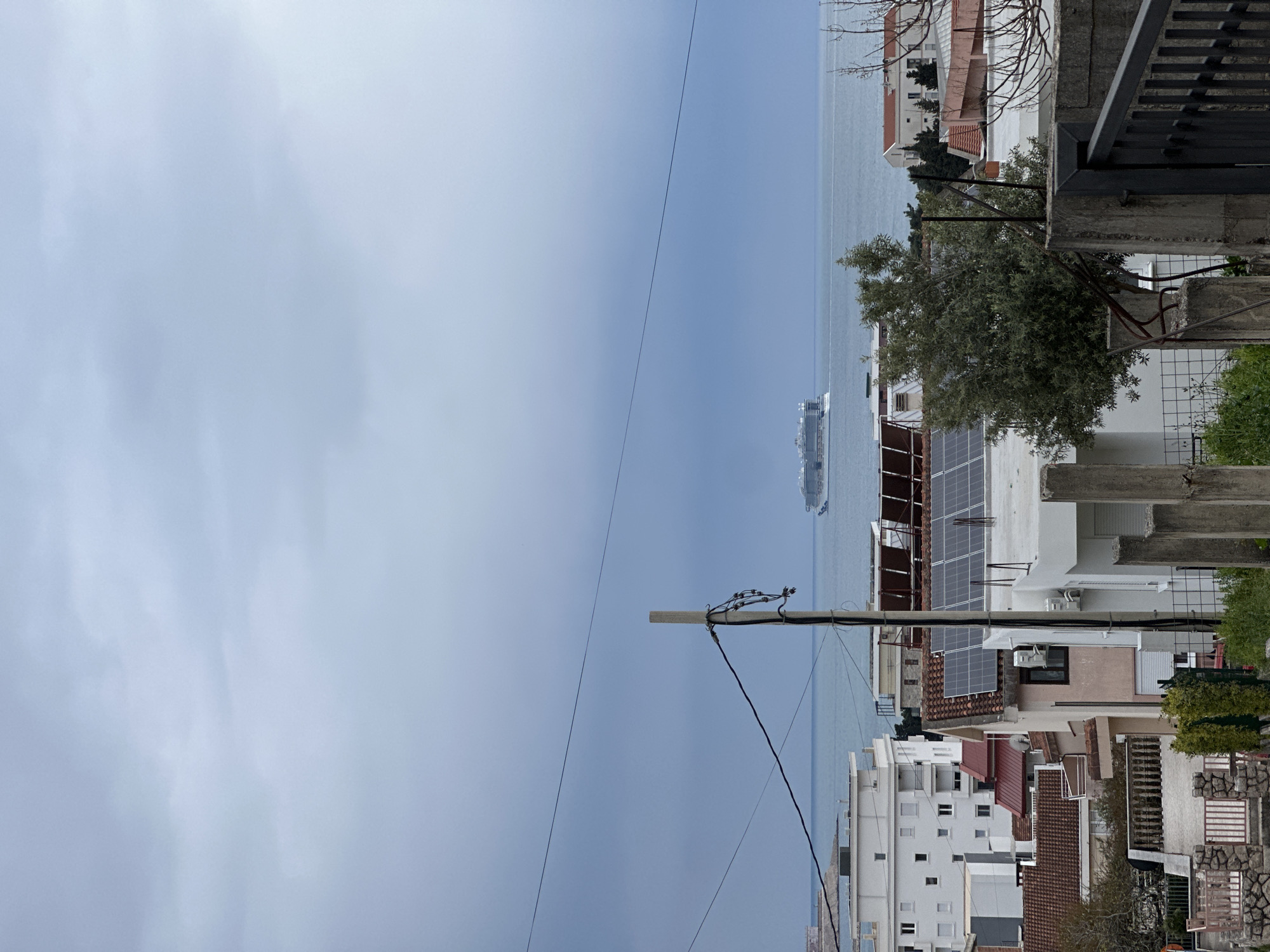 The scene is a view of the sea from what appears to be a neighborhood within Šušanj, Montenegro. A large cruise ship is visible on the water in the distance. In the foreground, there are residential buildings with solar panels on the roof of one building. A utility pole with power lines crosses the sky. The sky is overcast. The angle of the shot suggests it was taken from a high vantage point, possibly a balcony or an upper floor of a building.