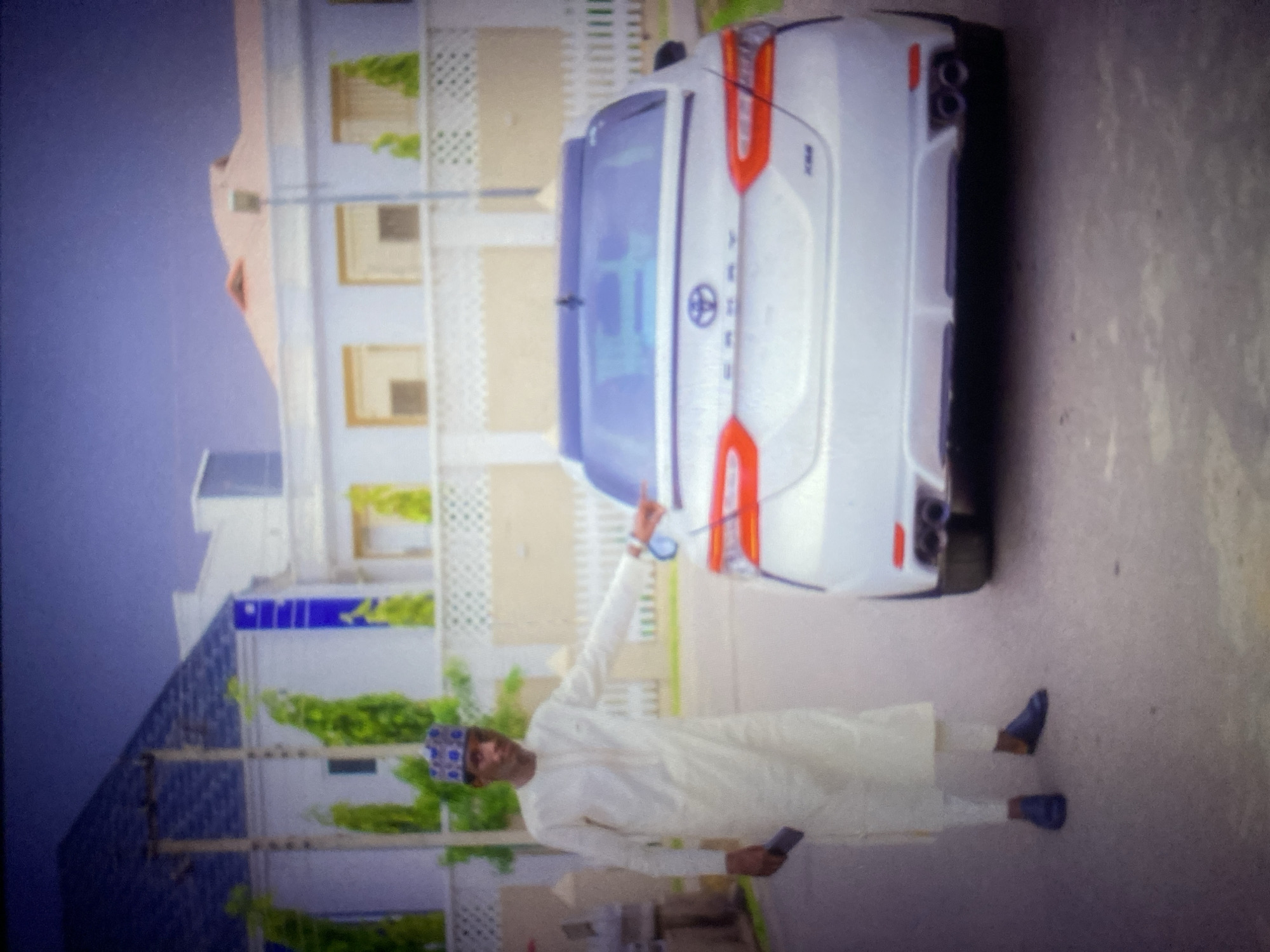 A man stands next to a white Toyota Camry on a paved street. The man is wearing a light-colored traditional outfit and a patterned cap. He is holding a dark object, possibly a phone, in his left hand and pointing at the car with his right hand. The car's rear is visible, showing its taillights and dual exhaust pipes. In the background, there are modern houses with white fences and lush green plants. The sky above is a deep purple. The overall impression is one of an affluent urban setting.