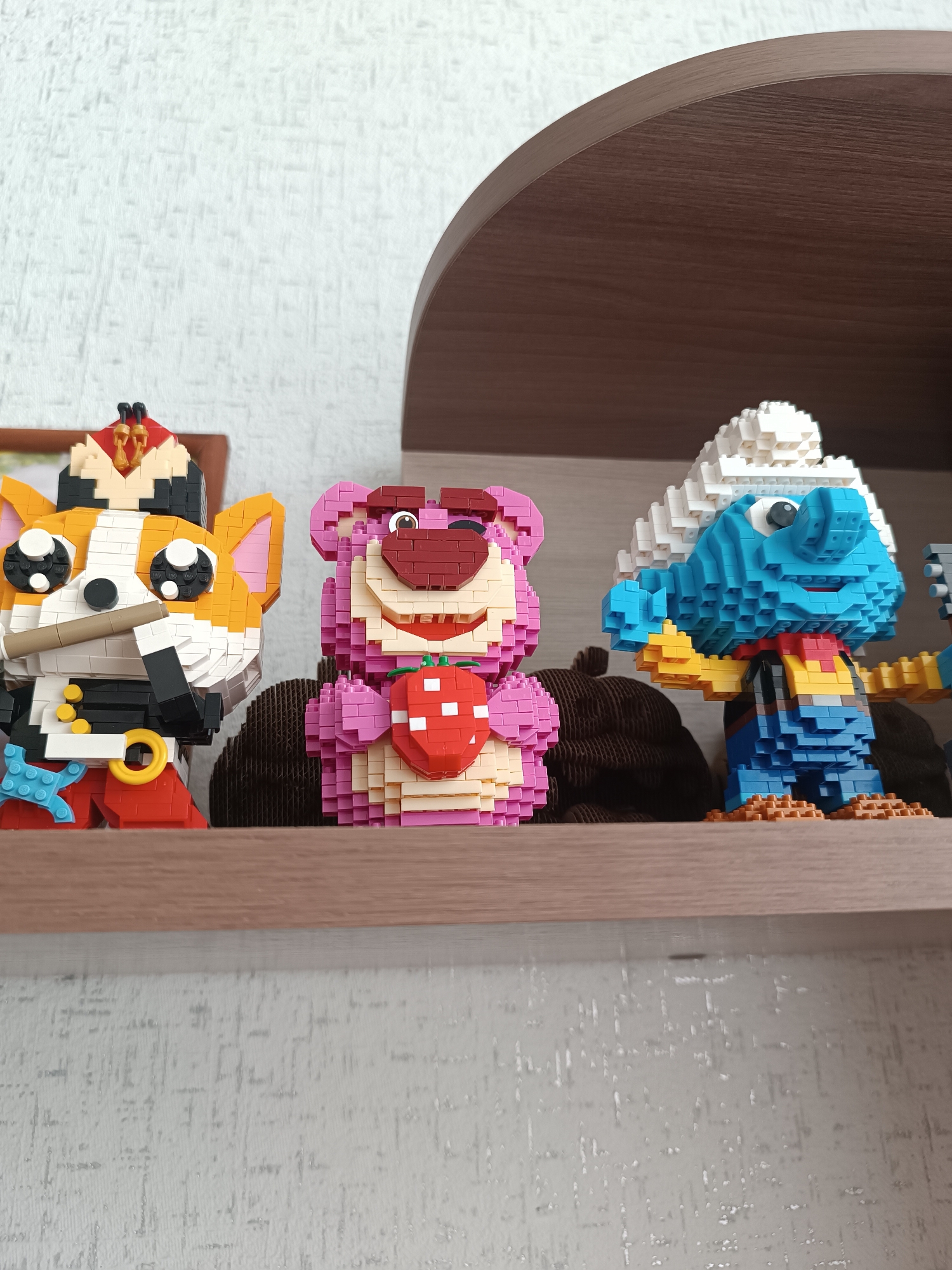 A shelf in an indoor environment holds three LEGO-like figures. From left to right, the figures are: a cartoon character resembling a cat in a military-style outfit with a hat and holding a tan-colored stick; a pink bear holding a red strawberry; and a blue Smurf-like character wearing a white hat and a yellow and red outfit with blue overalls. The shelf is made of brown wood and is attached to a wall with a textured, off-white wallpaper. There are some blurry objects behind the figures. This picture appears to be taken in the city of Chaikovsky, Russia.