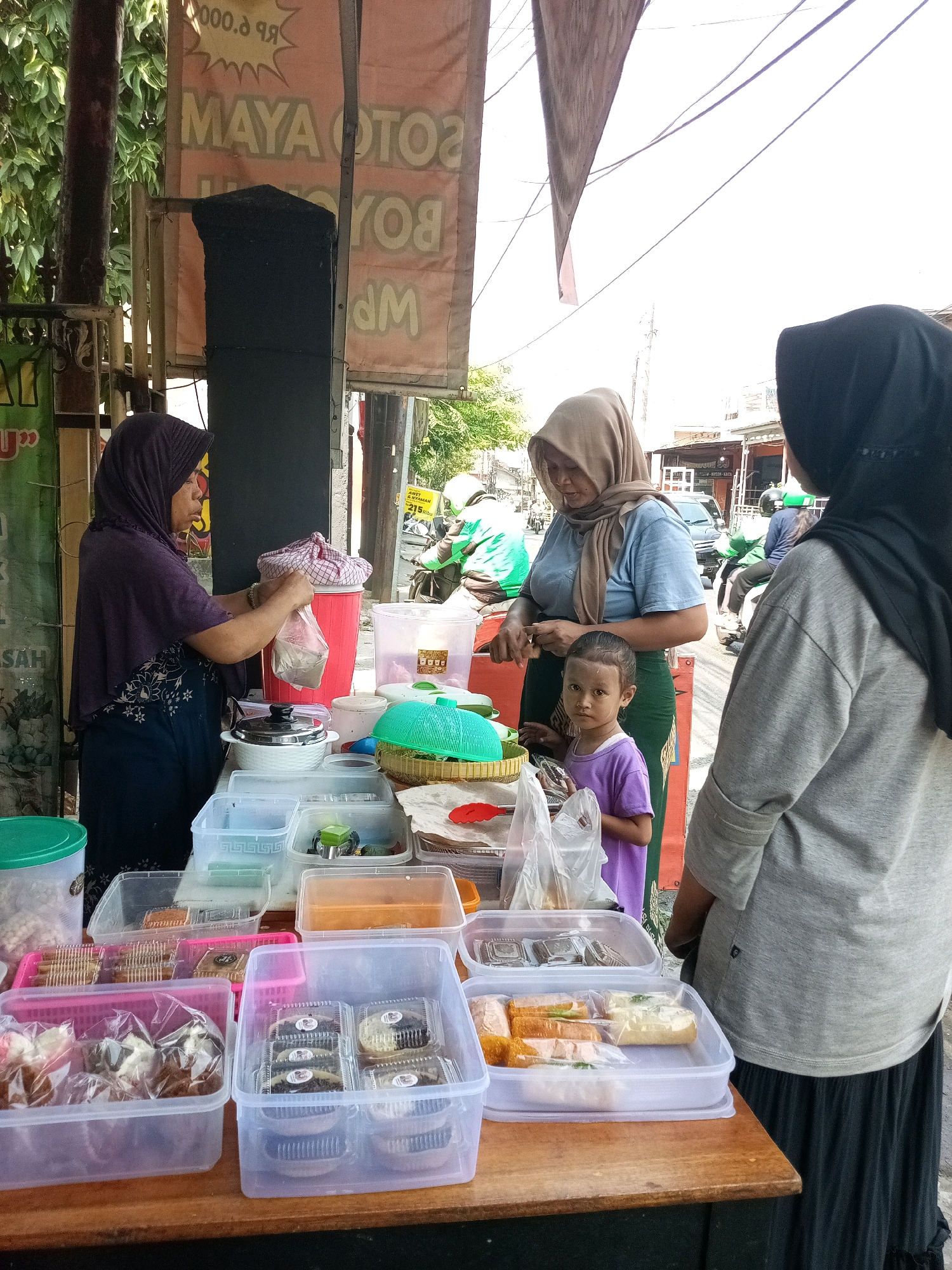 This is a medium-shot, eye-level, outdoor photograph of a food stall in Bekasi, Indonesia. In the foreground, a wooden table displays various food items in clear plastic containers. On the left, a woman wearing a dark purple hijab and a matching dress is serving food from a large red cylindrical container into a clear plastic bag. She is reaching into the container with both hands. Next to her, there are several clear plastic containers filled with what appear to be pastries or snacks, some packaged in smaller clear plastic bags.
Further back, a woman wearing a tan hijab and a blue shirt is standing behind the table, looking down at something. To her right, a young child in a purple t-shirt is standing and looking towards the camera. To the right of these two figures, another person wearing a black hijab and a grey long-sleeved shirt is facing away from the camera, looking towards the food stall.
In the background, there are more food stall elements, including a black speaker. Behind the stall, there is a street scene with buildings, a car, and several motorcycles, one of which has a rider in a green jacket. Trees with green foliage are visible on the left side of the image, and a bright, slightly overcast sky fills the upper portion of the frame. A banner with Indonesian text, including "MAYA SOTO BOYOLALI," is visible hanging behind the stall.