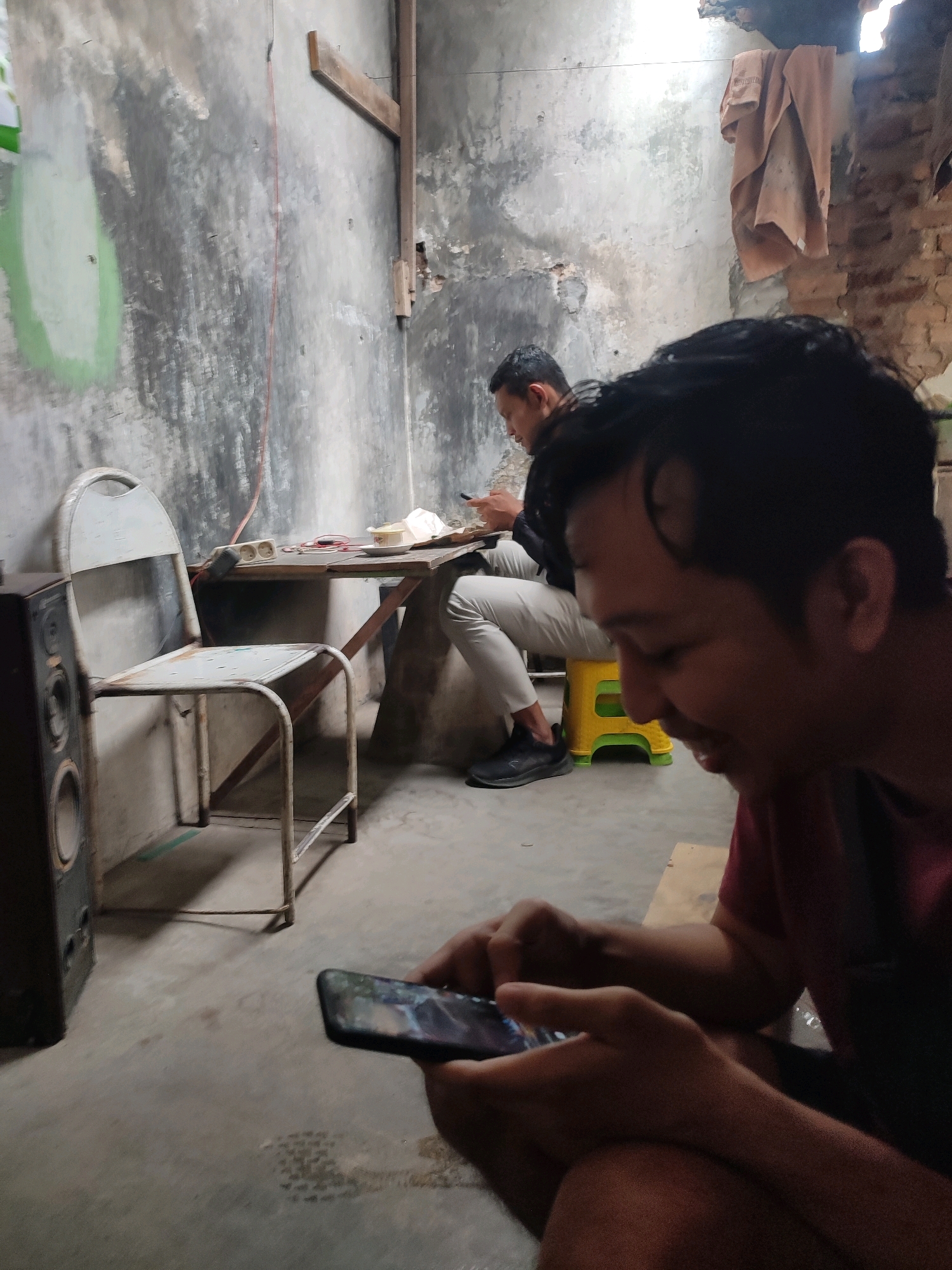 An indoor scene depicts two men in what appears to be a dimly lit, rustic dwelling in Trenggalek, Indonesia. In the foreground, a man wearing a maroon t-shirt is seated, looking down at a smartphone held in his hands. The phone screen displays a colorful image, possibly a game. His right hand is positioned on the screen, suggesting he is actively interacting with it. Behind him, another man is seated at a table, also engaged with his smartphone. He is wearing a black jacket and light-colored pants. The environment is characterized by worn, textured walls, a simple metal chair next to a speaker, and a makeshift table made of wood. A light-colored towel hangs from the ceiling in the background. The lighting suggests it is nighttime or the room has limited natural light.