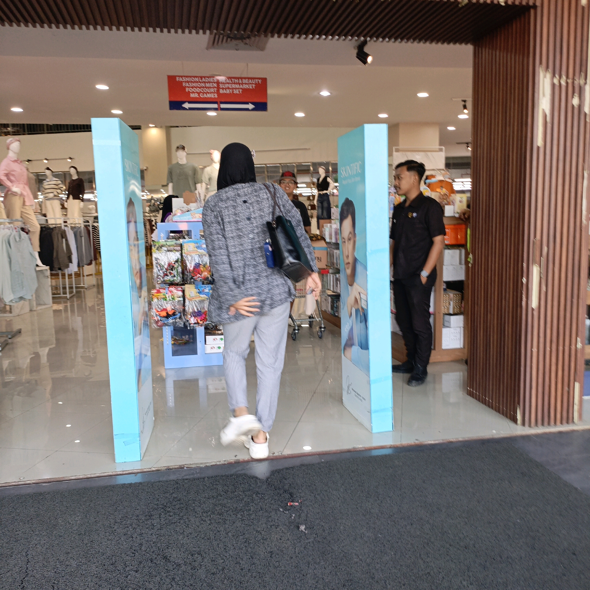 This image depicts the entrance of a retail establishment in Salatiga, Indonesia. The scene is viewed from a low angle looking towards the interior of the store. In the foreground, a dark grey textured mat is visible.
The central focus is a person wearing a gray patterned long-sleeved top, grey striped pants, and white sneakers, seen from the back as they walk through a doorway. They are carrying a dark handbag over their left shoulder. To the right of this individual, a uniformed male, likely security personnel, stands by a wooden slatted partition. He is wearing a black short-sleeved collared shirt and black pants, with a watch on his left wrist.
Flanking the entryway are two large, bright blue display panels. The panel on the left is mostly out of frame, while the panel on the right features a large printed image of a person's face, identified by the text "SKINTIFIC" above it. Behind the main figures, the store interior is visible, with clothing racks, mannequins, and shelving units displaying various products. A red and white directional sign hangs from the ceiling, indicating "FASHION LADIES", "HEALTH & BEAUTY", "FASHION MEN", "SUPERMARKET", "FOODCOURT", "BABY SET", and "MR. GAMES". The floor inside the store appears to be light-colored tile with a reflective surface. The lighting is artificial, with several ceiling lights visible.