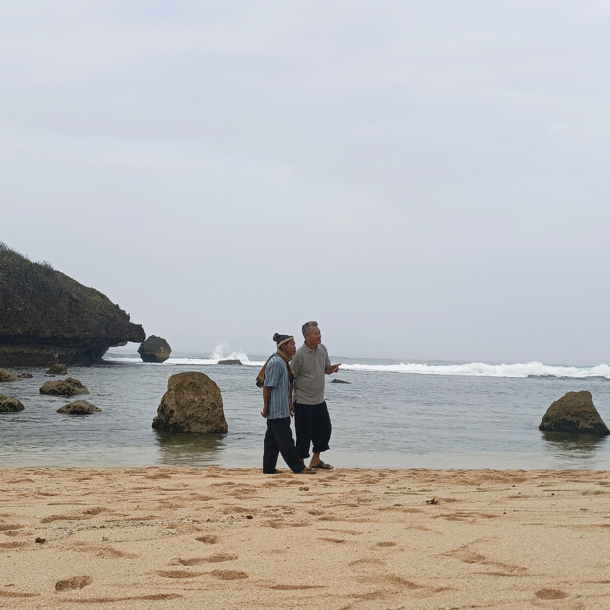 Two elderly men are walking on a sandy beach. The man on the left is wearing a blue and white striped long-sleeved shirt and black pants, with a patterned head covering and a scarf draped around his neck. The man on the right is wearing a light grey polo shirt and black pants. They are both looking towards the right side of the frame, with the man on the right pointing with his right index finger. The beach has a coarse, light brown sand with visible footprints and small shells. The sea is calm with gentle waves breaking in the distance. Several large, brown rocks are scattered in the water and on the beach. To the left, a large, moss-covered rock formation juts out into the sea, featuring an archway. The sky is overcast and grey. The scene appears to be located at a beach in Tepus, Indonesia.