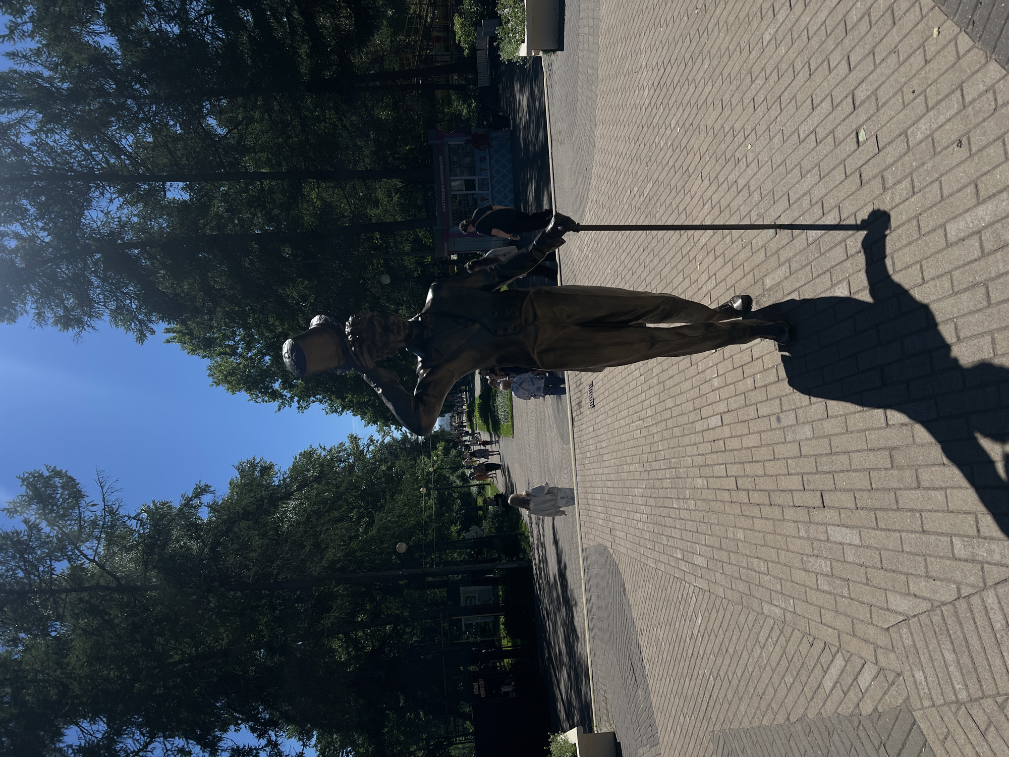 The image depicts a bronze statue of a man in a top hat and suit, holding a cane and balancing a bucket on his head. The statue is positioned on a brick-paved walkway in a park, with lush green trees lining the path and a clear blue sky overhead. Several people are visible in the background, walking along the path and near small kiosks. One kiosk has text in Russian that appears to advertise ice cream. The scene suggests a sunny day in a public recreational area, likely a park in Zelenogorsk, Russia, given the presence of Russian text and the typical appearance of such parks in the region. The statue casts a long shadow on the patterned brickwork.