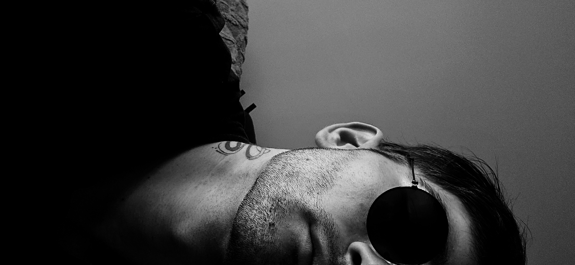 The image is a black and white, low-angle portrait of a person, likely male, captured from the right side of their head and upper torso. The subject is oriented vertically within the frame, suggesting they are lying down or leaning back with the camera positioned below and tilted. The person has short dark hair and a short beard or stubble. They are wearing dark, round sunglasses that entirely cover their right eye, obscuring any discernible expression.
Tattoos featuring stylized circular or eye-like designs are visible on the subject's neck and upper shoulder area. The person is dressed in dark clothing, possibly a t-shirt or hoodie, which is mostly engulfed in deep shadow on the left side of the image. A textured surface, perhaps a wall or piece of furniture, is faintly visible behind the left shoulder/head. The background is a plain, light grey wall, providing a stark contrast to the dark tones of the subject's clothing and accessories.
The lighting creates a dramatic effect with strong contrasts between highlights on the face and ear, and deep shadows across the left side of the frame. The black and white aesthetic enhances the artistic and somewhat contemplative mood of the photograph. No specific activity is taking place beyond posing or resting, and no text is visible in the image. The setting is an indoor location, with the given context being Plovdiv, Bulgaria.