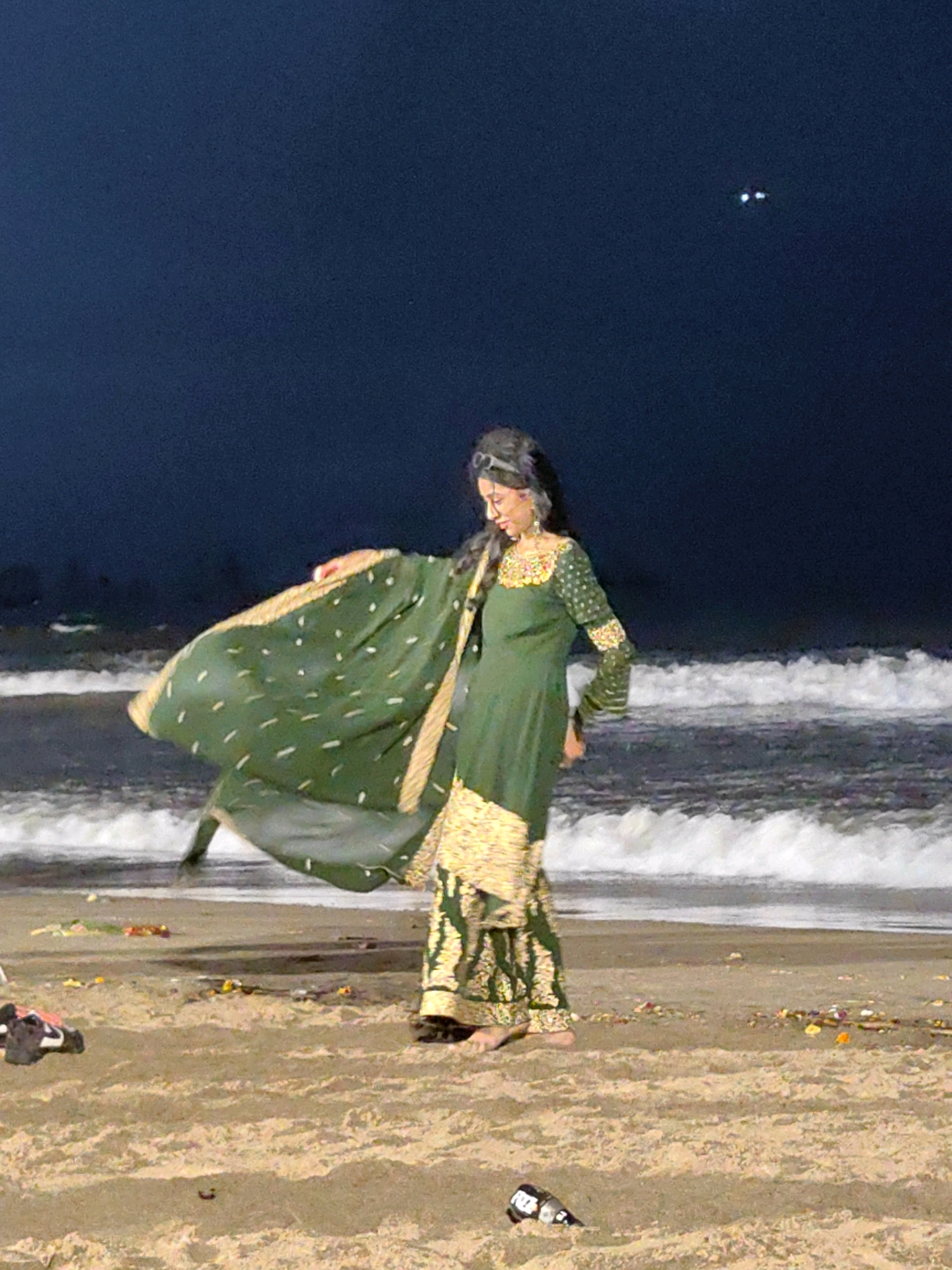 A young woman is standing on a beach at night. She is wearing a traditional green and gold Indian outfit. The outfit consists of a long-sleeved dress with intricate gold embroidery around the neckline and cuffs, and a matching wide-legged pant or skirt. She is also holding a large, flowing green dupatta with gold accents. The dupatta is draped over her arm and extends out to her left, creating a dynamic visual effect. The woman is looking down and to her left, with her right hand gently touching the dupatta.
The beach sand is visible in the foreground, with some debris scattered on it, including a black and white bottle and some dark clothing items. In the background, waves are crashing onto the shore. The ocean is dark, and the sky above is a deep navy blue with two bright, distinct lights, possibly from a distant boat or aircraft. The overall lighting suggests it is nighttime, with ambient light from the sky and possibly some artificial light sources not visible in the frame illuminating the scene. The setting appears to be a beach in Mumbai, India, given the common attire and typical beach scene at night.