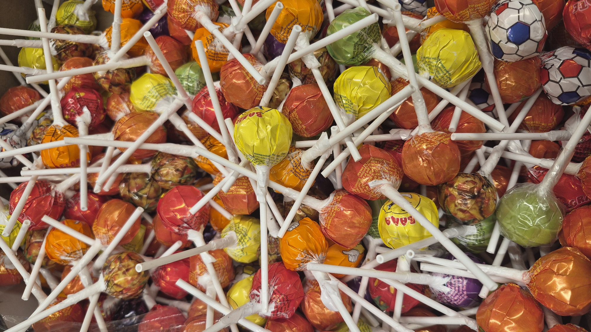 The image shows a close-up, overhead view of a large pile of individually wrapped lollipops. The lollipops have brightly colored wrappers, including orange, red, yellow, green, and purple. Many of the wrappers have graphic designs, such as smiley faces, fruit patterns, and what appear to be soccer balls. The lollipops are arranged haphazardly, with white plastic sticks protruding in various directions. There are no people or animals visible in the image. The lighting appears to be artificial and even, suggesting an indoor setting. No specific location in Dagda, Latvia can be determined from the visual information provided.