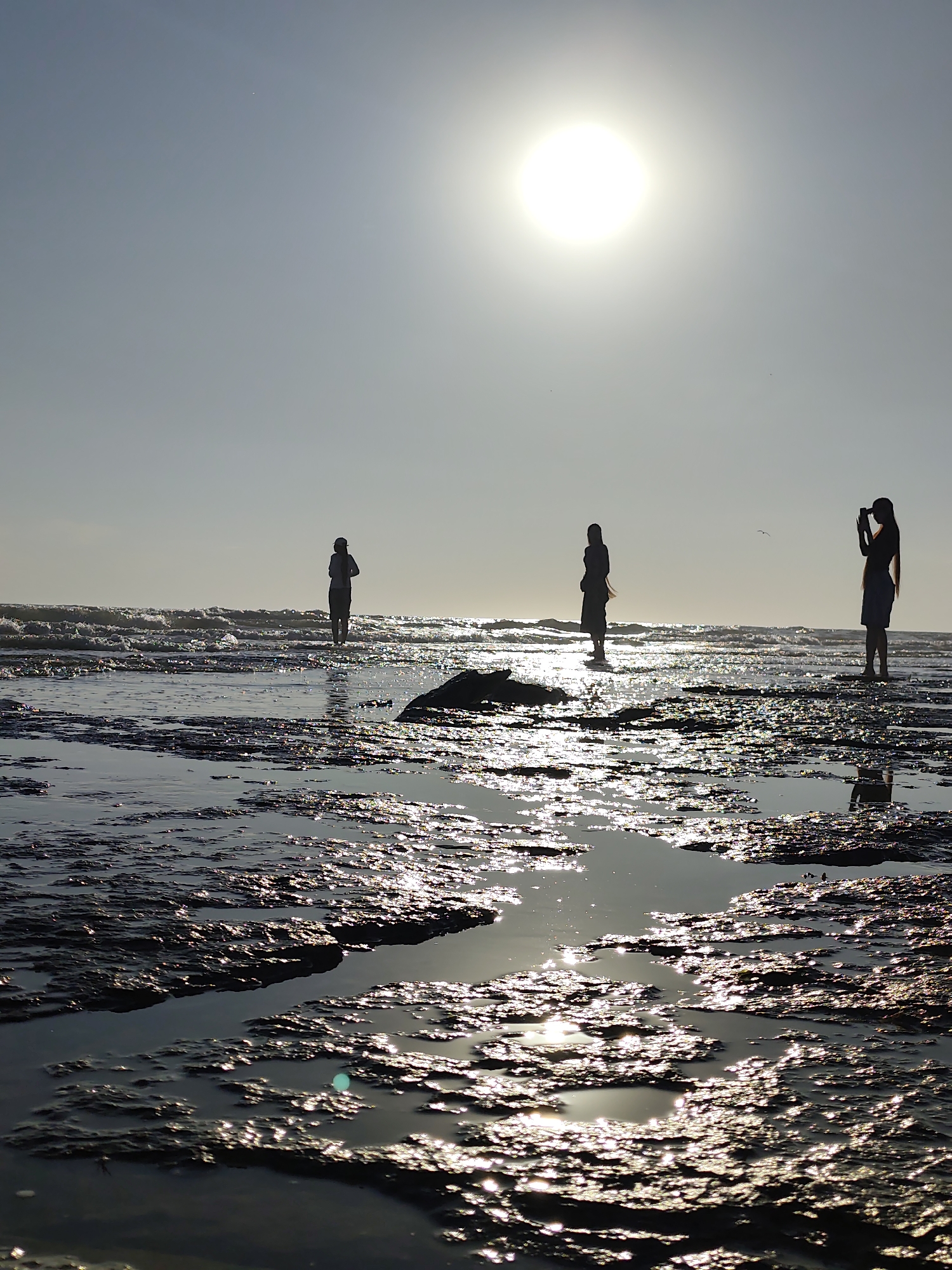 Three silhouetted figures stand on a rocky shoreline under a bright, hazy sun. The sun, a brilliant disc, is positioned high in the sky. The figures are spaced apart, with one on the far left seemingly observing the water, a second in the center looking down or holding something, and the figure on the far right holding a phone up as if taking a photograph. The foreground consists of shallow pools of water reflecting the sunlight, interspersed with dark, textured rocks. The middle ground shows the ocean with waves breaking, and the background is a hazy sky. This scene is consistent with the shores of the Caspian Sea near Aktau, Kazakhstan, known for its unique rocky coastlines and reflective waters, especially during sunrise or sunset.