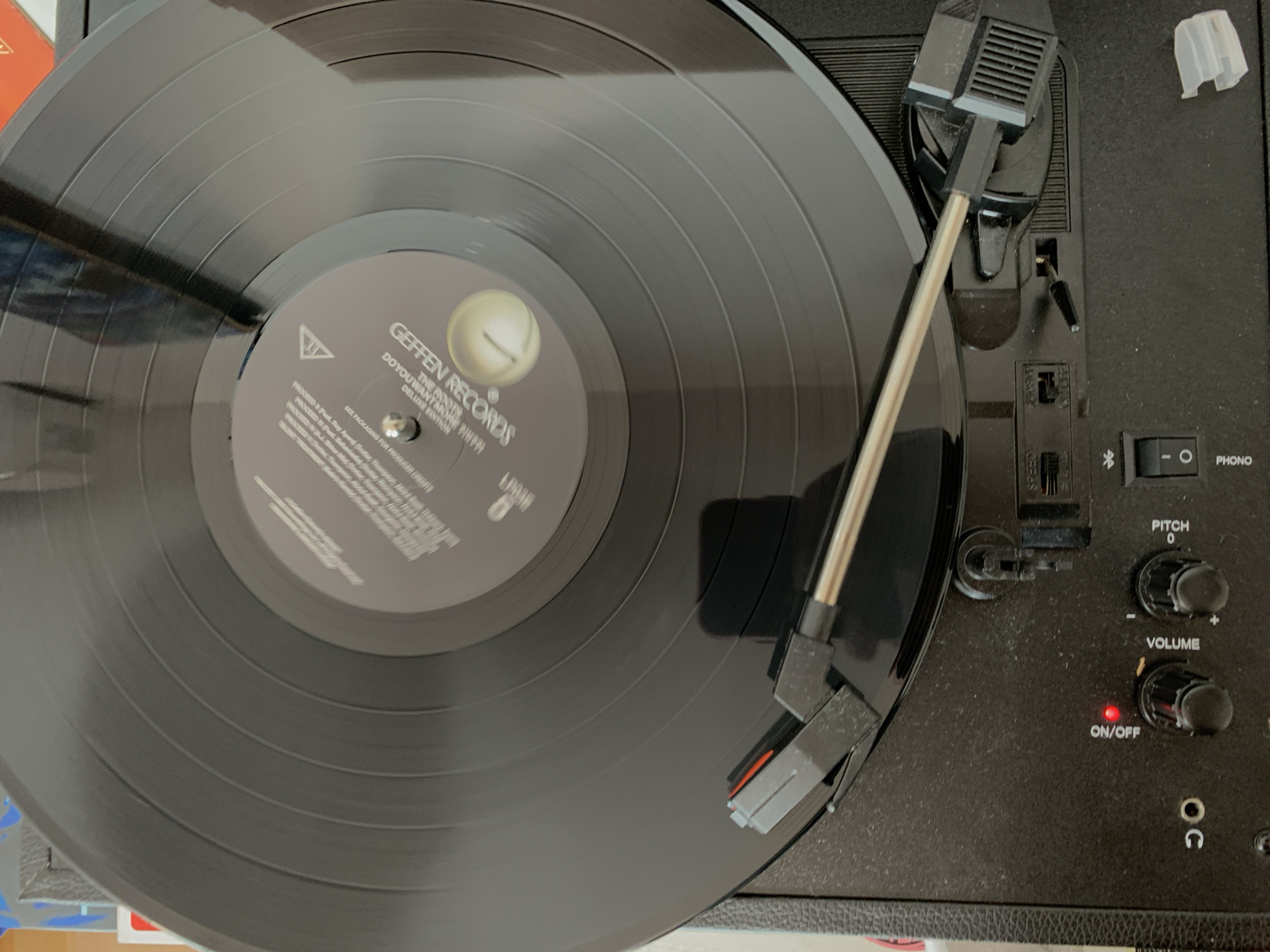 A black vinyl record is spinning on a turntable. The record has grooves that are visible, and there is a label in the center with text that is difficult to read. A tonearm with a needle is positioned over the record. The turntable is black and has several knobs and switches on its surface. A red light is illuminated on the turntable, indicating it is on. The background is out of focus and appears to be a room. There is no indication of the location being Barcelona, Spain.