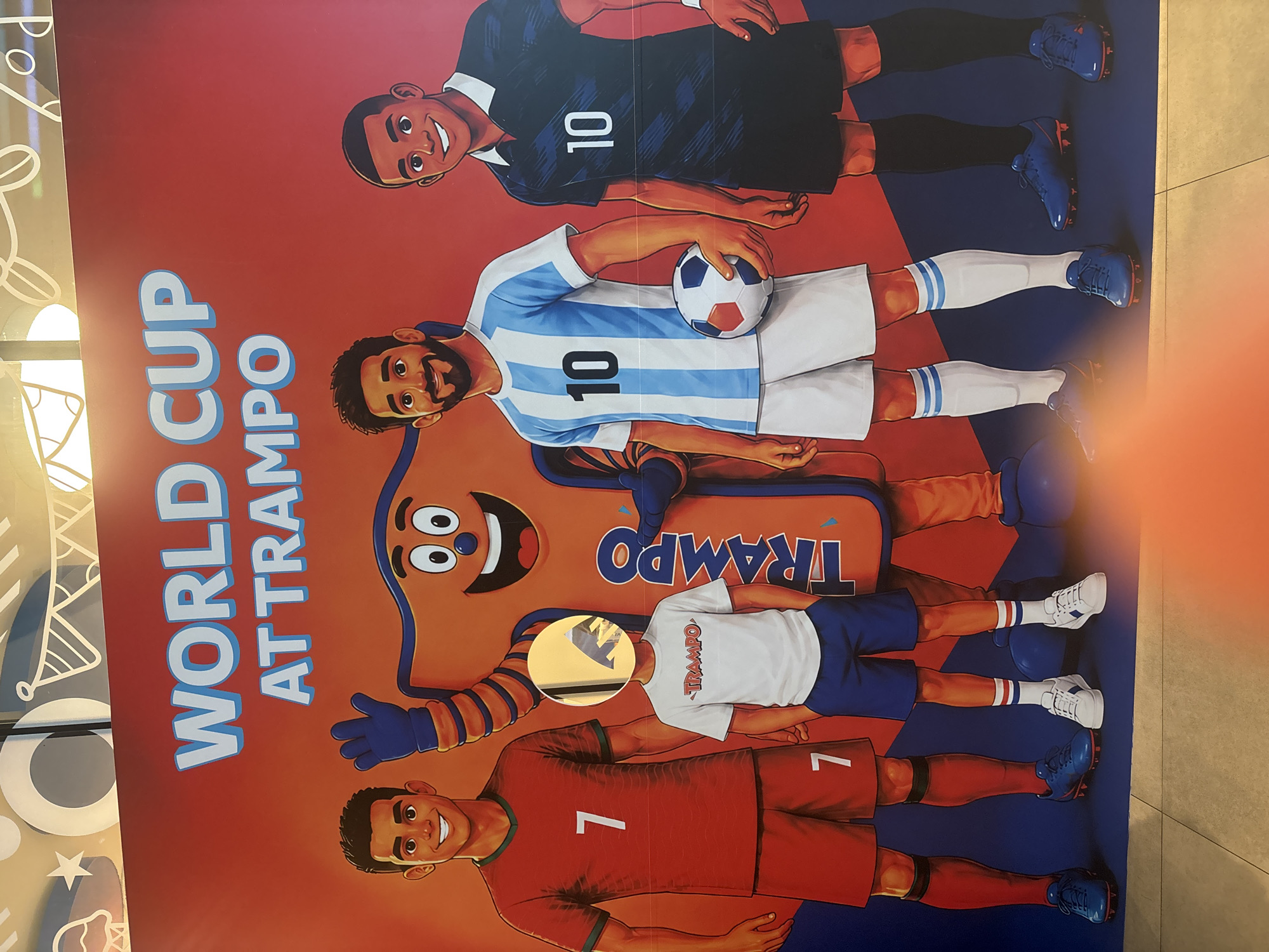 The image shows a life-sized promotional display for "World Cup at Trampo," likely a sporting event or activity at a venue called Trampo. The display features cartoonish representations of famous soccer players Cristiano Ronaldo (wearing a red jersey with the number 7) and Lionel Messi (wearing an Argentinian-style striped jersey with the number 10), alongside a referee-like figure (wearing a black jersey with the number 10). A mascot, an orange, amorphous character with a smiley face and blue gloves, stands between the players. In front of the mascot and between the two adult figures, a child stands wearing a white "TRAMPO" t-shirt and blue shorts, with their face obscured by a circular yellow sticker featuring a black and white dinosaur silhouette. The background is a vibrant orange banner with white lettering. The display appears to be located in a public space with a tiled floor and a textured ceiling, possibly a mall or entertainment complex. The lighting suggests an indoor environment. The scene is static and posed for a photograph.