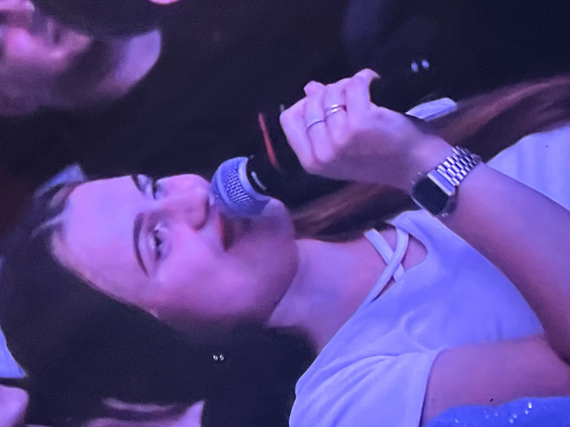 A young woman with dark hair and light skin, wearing a white t-shirt and a watch on her left wrist, holds a microphone to her mouth. Her eyes are directed to her right, and she has red lipstick on. She is wearing two rings on her right hand. The scene is dimly lit with purple and pink lighting. This image was taken in the city of Genemuiden, Netherlands.