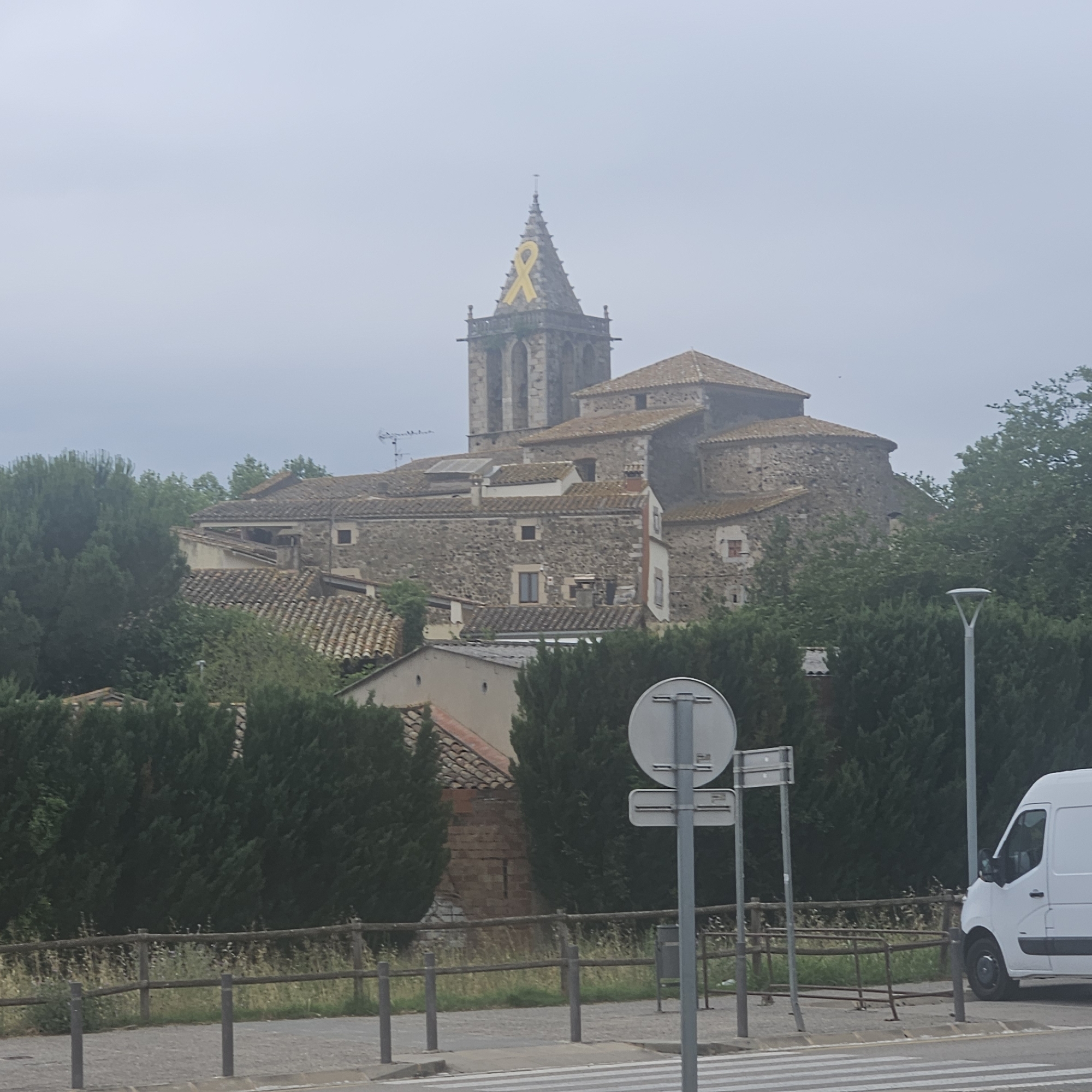 The image shows a stone church with a tall, pointed steeple adorned with a large yellow ribbon. The church is surrounded by trees and other buildings with terracotta tile roofs. In the foreground, there is a white van, a lamppost, a traffic sign, a wooden fence, and a pedestrian crossing. The sky is overcast, creating a muted light over the scene. The church appears to be located in a village or small town, possibly in Aiguaviva, Spain, given the presence of Spanish architectural styles and the yellow ribbon, which is a symbol of political protest in Catalonia.