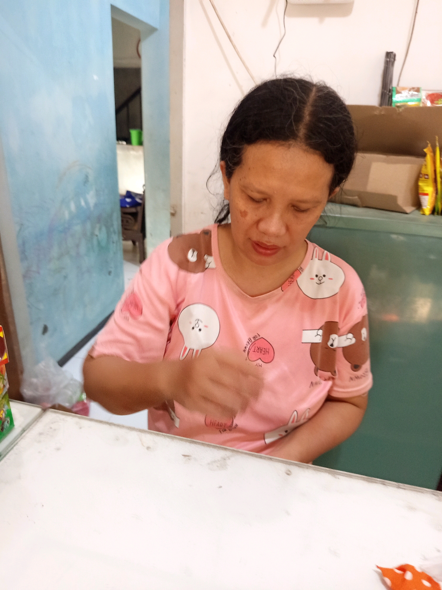 The image shows a woman with dark hair and brown skin sitting at a white table. She is wearing a pink t-shirt with cartoon characters on it. Her eyes are closed and her head is bowed. Her right hand is blurred in motion, suggesting she is either reaching for something or gesturing. The background is out of focus but appears to be an indoor setting. To the left, there is a light blue wall with some graffiti or drawings on it. To the right, there is a green cabinet or refrigerator and some shelves with items on them. A white electrical cord hangs from the ceiling. A corner of a window or doorway is visible between the blue wall and the white wall. The overall impression is of a casual, domestic environment in Singosari, Indonesia.