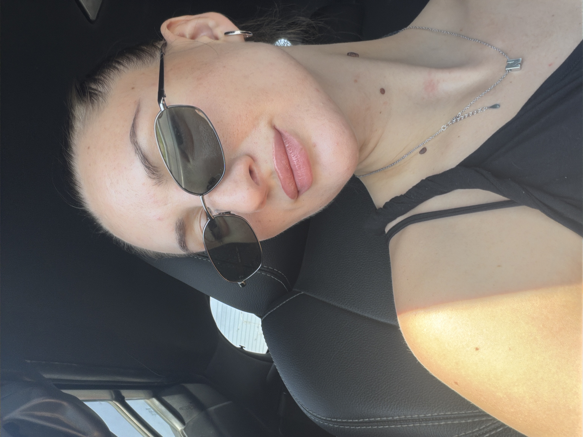 A young woman wearing sunglasses and a black top is pictured inside a car. The car appears to be parked, as indicated by the stillness of the subjects and the static background. The woman is looking towards the camera, with her head slightly tilted. She has her hair pulled back and is wearing a silver necklace with a pendant. The background shows the interior of the car, including a black leather seat, and the exterior through a window, revealing a bright, sunny day. Based on the typical weather patterns and the presence of cars and modern infrastructure, this scene could plausibly be located in a city like Sharm El Sheikh, Egypt, known for its sunny climate and tourist resorts.
