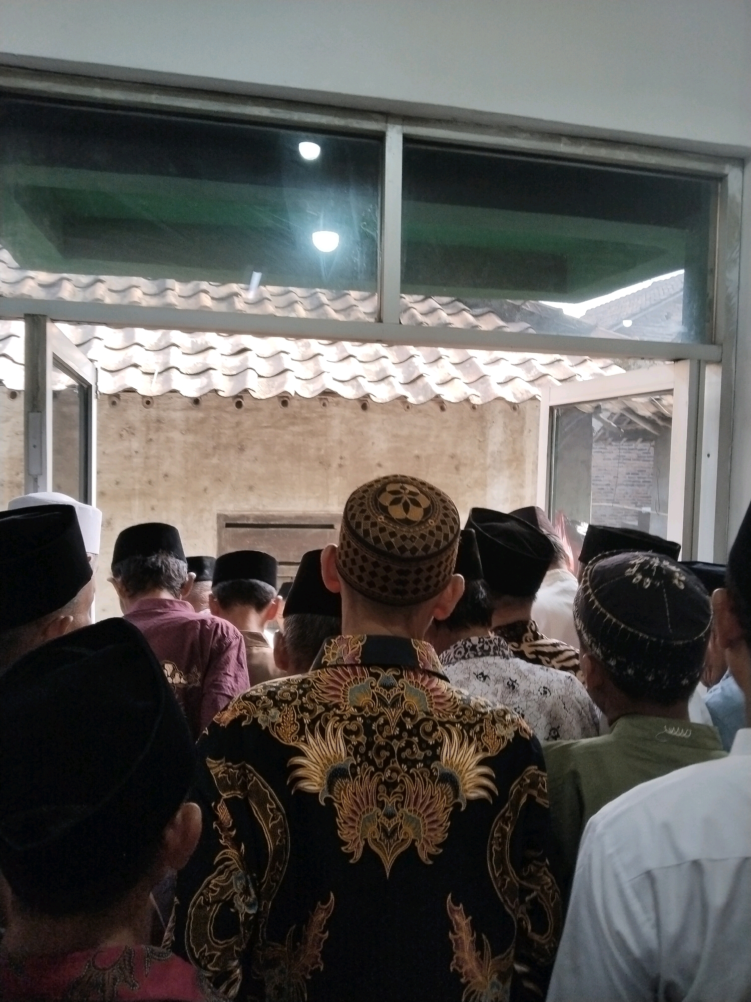 A group of men wearing traditional Muslim attire, including peci hats and patterned shirts, are gathered in what appears to be an outdoor courtyard or gathering space. The men are facing away from the camera, suggesting they are participating in a communal activity such as a prayer or ceremony. In the background, a tiled roof and a light-colored wall are visible. Above the roofline, a large window with reflective glass shows a glimpse of an interior space with what seem to be lights and possibly a green ceiling. The overall scene suggests a cultural or religious gathering in a location that combines outdoor and indoor elements. The presence of peci hats and the style of clothing suggest a setting in Indonesia, potentially within the city of Serang, known for its significant Muslim population and cultural heritage.