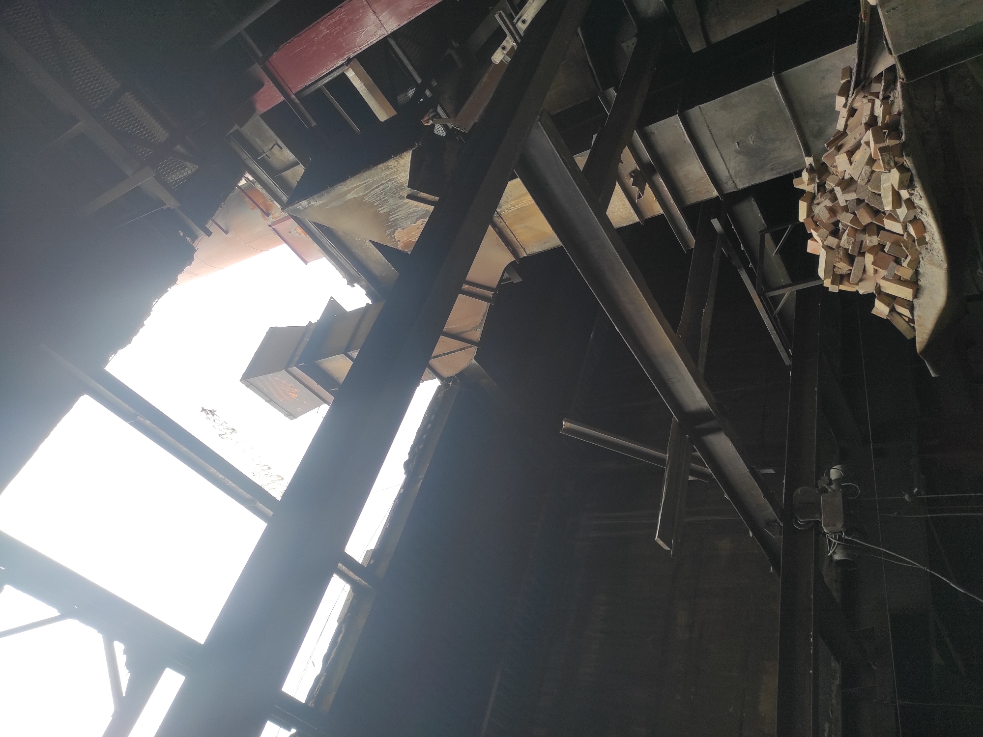 The image depicts the interior of a derelict industrial building, likely a factory or power plant, located in the city of Kyzyl, Kazakhstan. The structure shows significant disrepair, with a partially collapsed roof allowing bright daylight to filter through. Large, dark metal beams crisscross the space, supporting the remains of the upper levels. Various industrial components, including large ventilation ducts and metal structures, are visible throughout the scene. In the foreground, a pile of orange bricks rests on a metallic surface, suggesting recent demolition or ongoing disuse. The overall environment is dark, dusty, and industrial, characterized by decay and abandonment. There are no people present in the image.