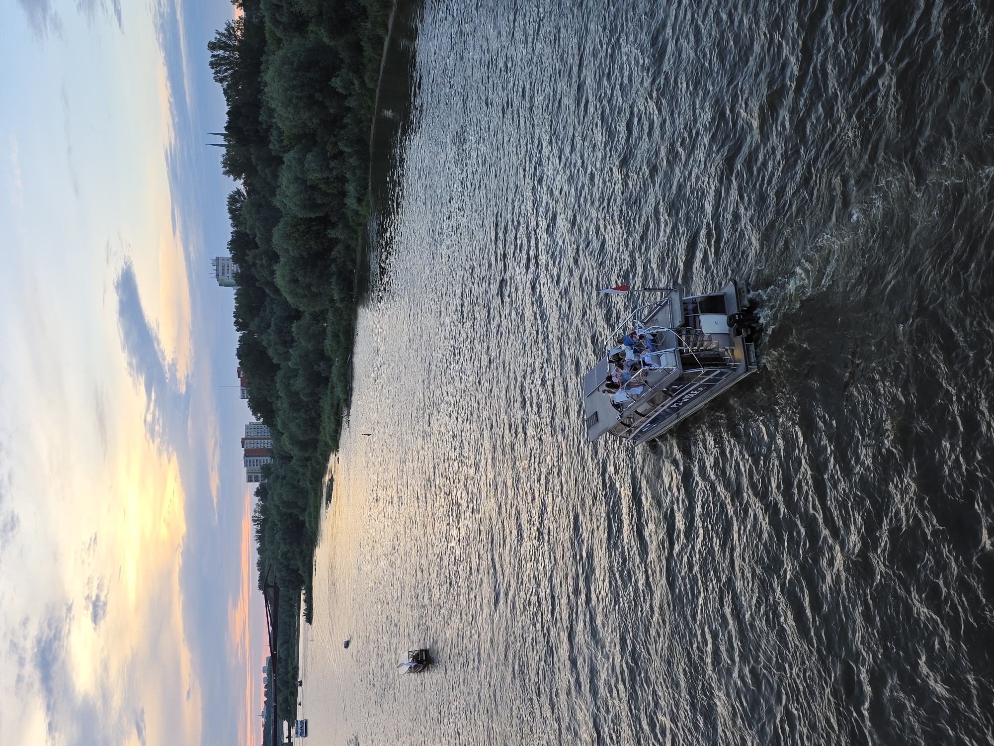 The image captures a scenic view of the Vistula River in Warsaw, Poland, during what appears to be sunset. Several boats are visible on the water. In the foreground, a larger tour boat carrying passengers is moving away from the viewer, creating a wake. The boat has a covered upper deck and an open lower deck where passengers are seated. A small Polish flag is attached to the stern of this boat.
Further in the distance, two smaller boats are also on the river. One is closer to the shore and appears to be a smaller tour boat with a white canopy and a flag. Another very small boat is further out.
The riverbanks are lined with lush green trees, and beyond the trees, city buildings are visible, including apartment blocks and what appear to be church spires. A red bridge structure can be seen in the far left of the image. The sky is painted with the warm hues of sunset, with soft oranges, yellows, and purples blending with the blue sky and scattered clouds. The water's surface is rippled, reflecting the colors of the sky.