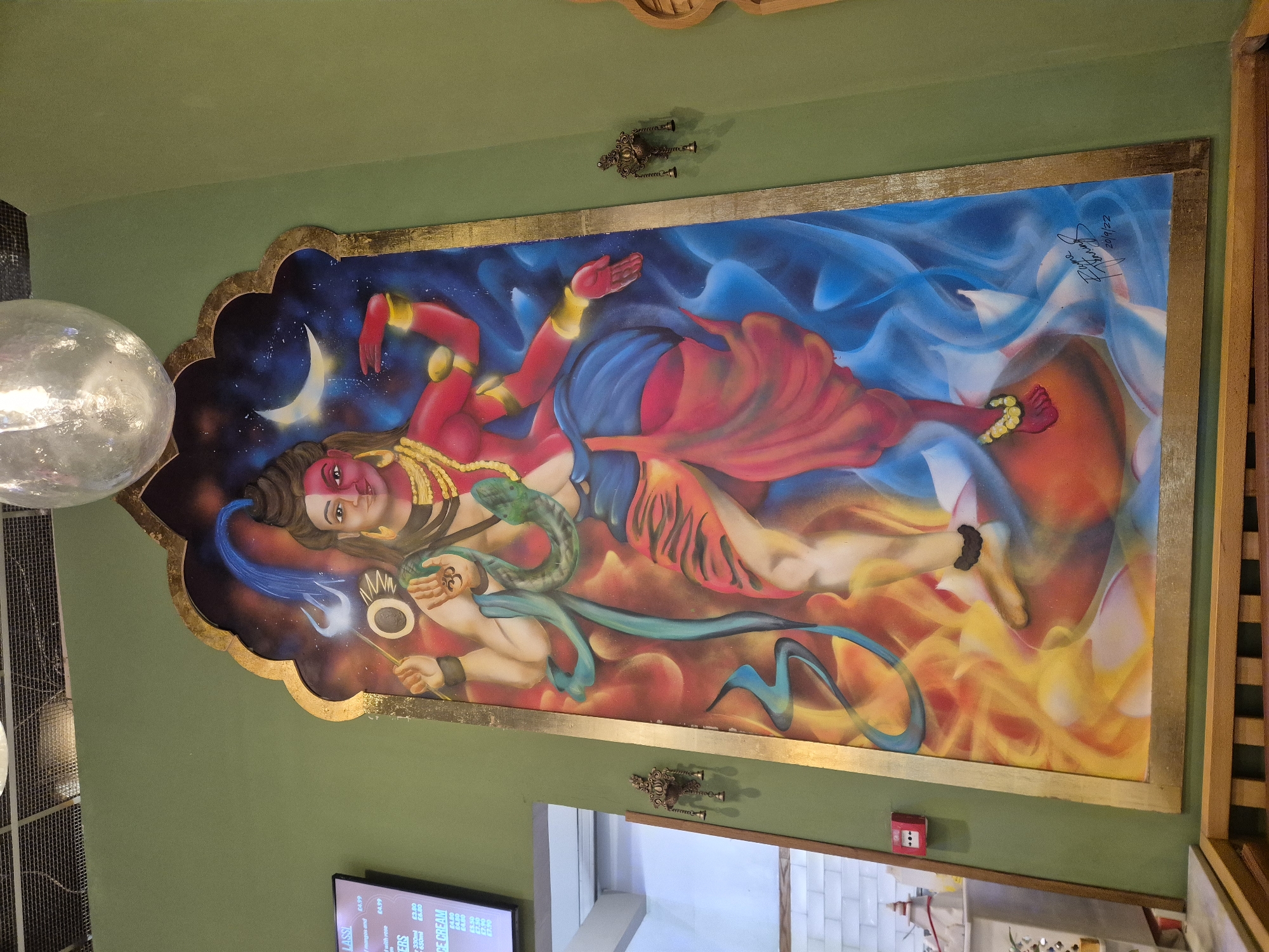 The image shows a painting on a wall, likely indoors in a commercial establishment. The painting is framed in gold and depicts a Hindu deity, possibly Shiva, lying down. The deity is multi-armed and has a snake draped around their neck. The background of the painting is divided into two parts: one side depicting a starry night with a crescent moon, and the other side with swirling blue and white smoke-like patterns. Beneath the deity are swirling patterns of orange and yellow. The painting is signed and dated "Jayne Fanning 20/9/22".
Two ornate, antique-style wall sconces are placed above and below the painting. The wall is painted a muted green color.
To the bottom left is a partial view of a light fixture and a menu screen, possibly displaying prices for food items such as "LASSI" and "ICE CREAM." A small red alarm pull box is seen on the wall below the painting. A glimpse of a white-tiled interior is visible in the bottom center, suggesting a kitchen or service area.
The location of the scene in the city of London, United Kingdom, is not stated and cannot be inferred from the image.