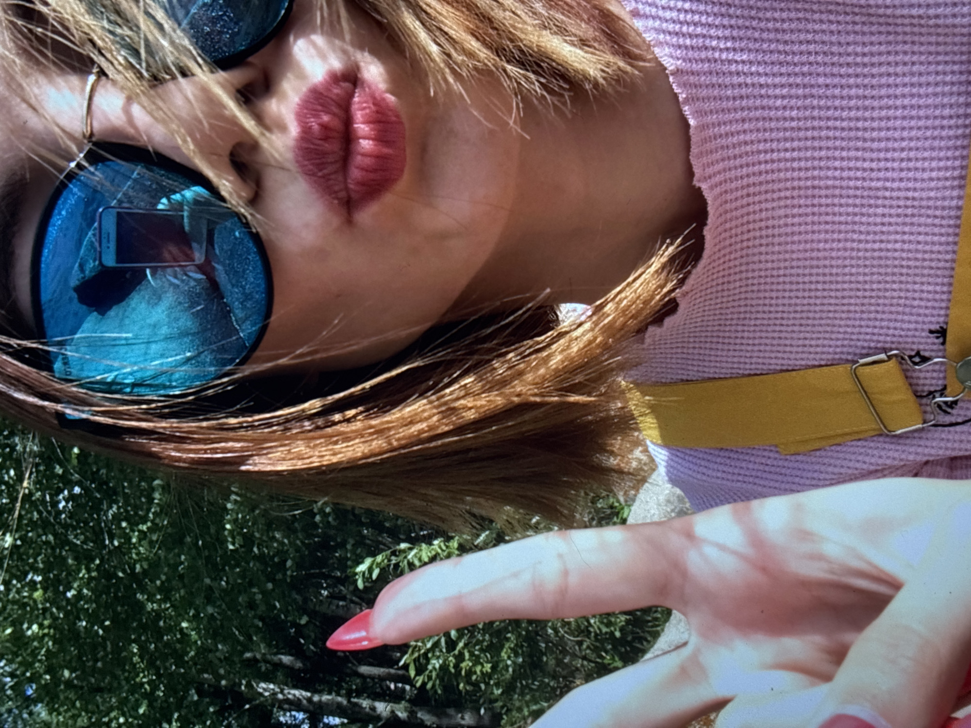 A close-up, low-angle selfie shows a person in an outdoor setting with trees in the background. The person is wearing sunglasses with bright blue lenses that reflect a mobile phone. They are also wearing a pink waffle-knit shirt and yellow straps. The person's lips are pursed, and their right hand is raised in a peace sign, with a pointed red nail visible. The lighting suggests it is daytime. The image does not contain any identifiable landmarks or features that would indicate it was taken in Omsk, Russia.