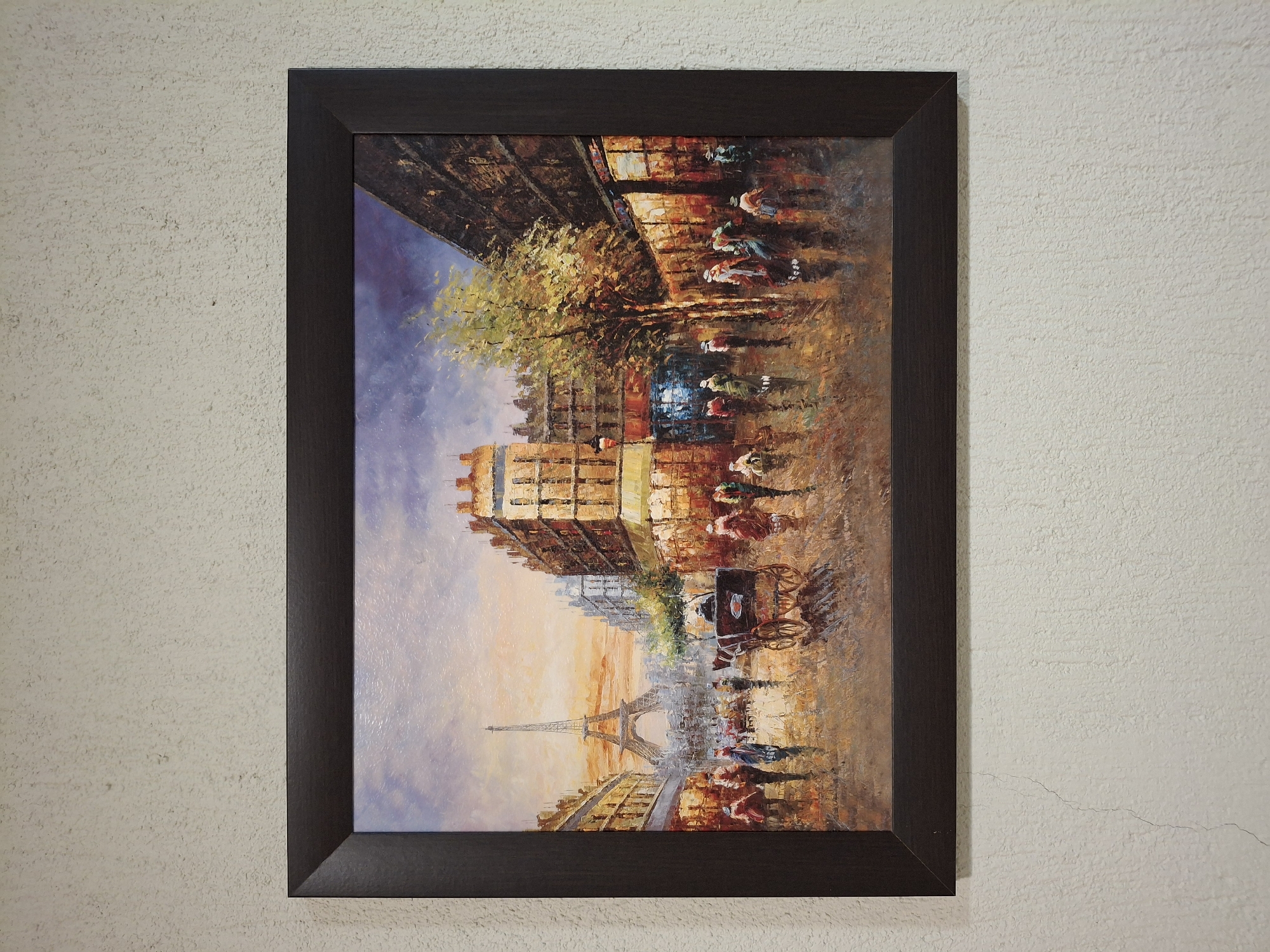 The image is a framed painting of a Parisian street scene featuring the Eiffel Tower in the distance. The painting depicts a street with buildings on either side, trees, and people walking. A horse-drawn carriage is also visible. The painting is in a dark brown frame and is hanging on a textured, off-white wall. The location is not in Krumovgrad, Bulgaria.