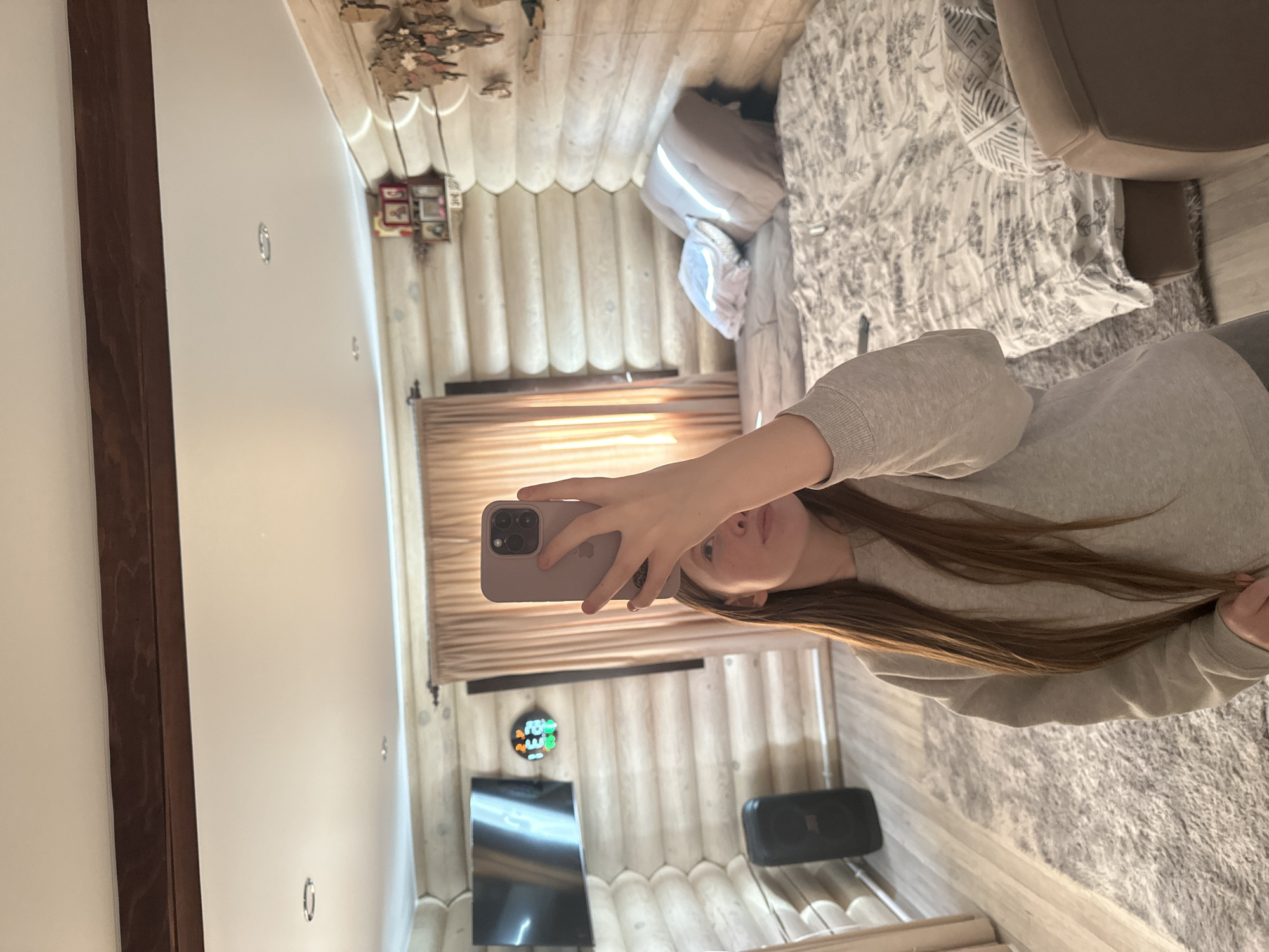 A person is taking a selfie in what appears to be a log cabin. The person is holding a purple smartphone, partially obscuring their face. They are wearing a grey sweater. The cabin walls are constructed from logs. A television, speaker, and a decorative clock are visible on one of the walls. A bed with patterned bedding and a chair are in the background. The location may be Ольховка, Russia, based on the request, although this cannot be definitively determined from the image alone.