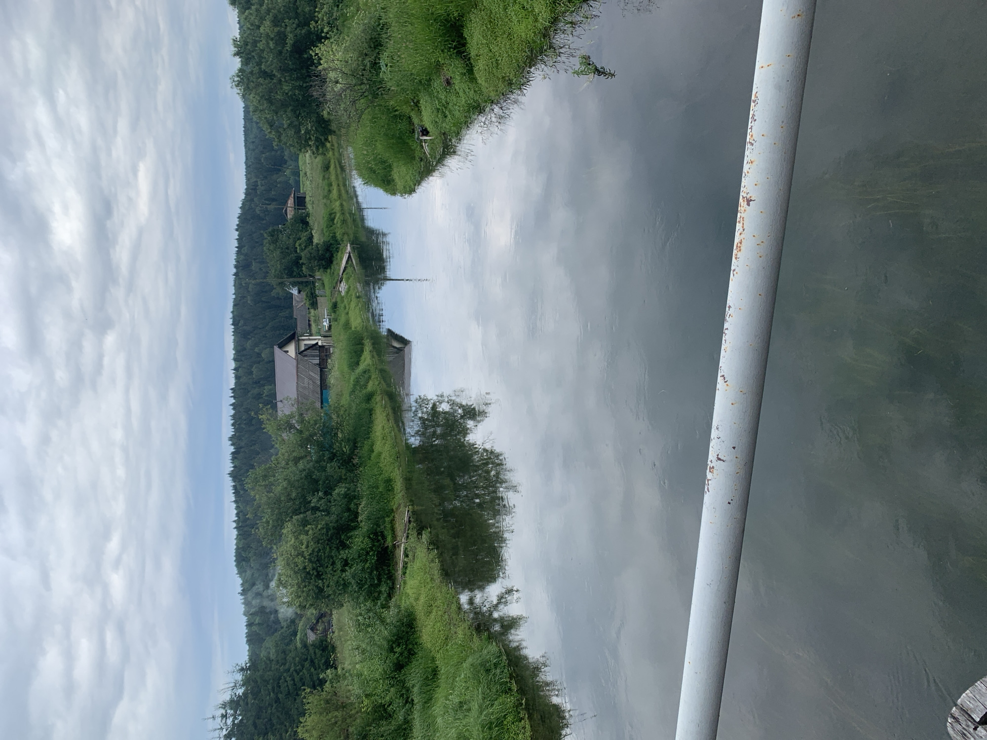 The image depicts a serene natural landscape with a river flowing through a rural area. The river is calm and reflects the cloudy sky and surrounding trees. On the left bank, lush green grass and trees grow, with a few houses nestled among them. Smoke is visible rising from one of the houses, suggesting activity. The right bank also features dense vegetation. In the distance, a thick forest stretches across the horizon under a heavily overcast sky. A grey, rusty metal railing runs across the bottom foreground of the image, possibly from a bridge or viewing platform. The scene appears to be in the city of Barsai, Russia, given the architectural style of the houses and the natural environment.