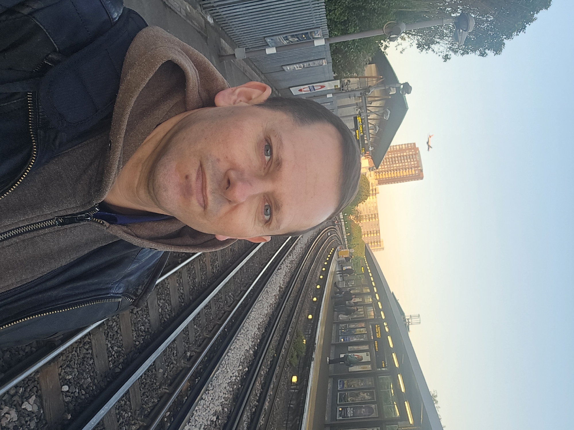 A man in a zipped, hooded jacket takes a selfie at a London Underground station, identifiable by the iconic red and blue roundel symbol. The photograph's viewpoint is from the trackside, with railway lines visible at the bottom left. People stand and wait on the platform. A building rises in the background, with a plane visible in the sky.