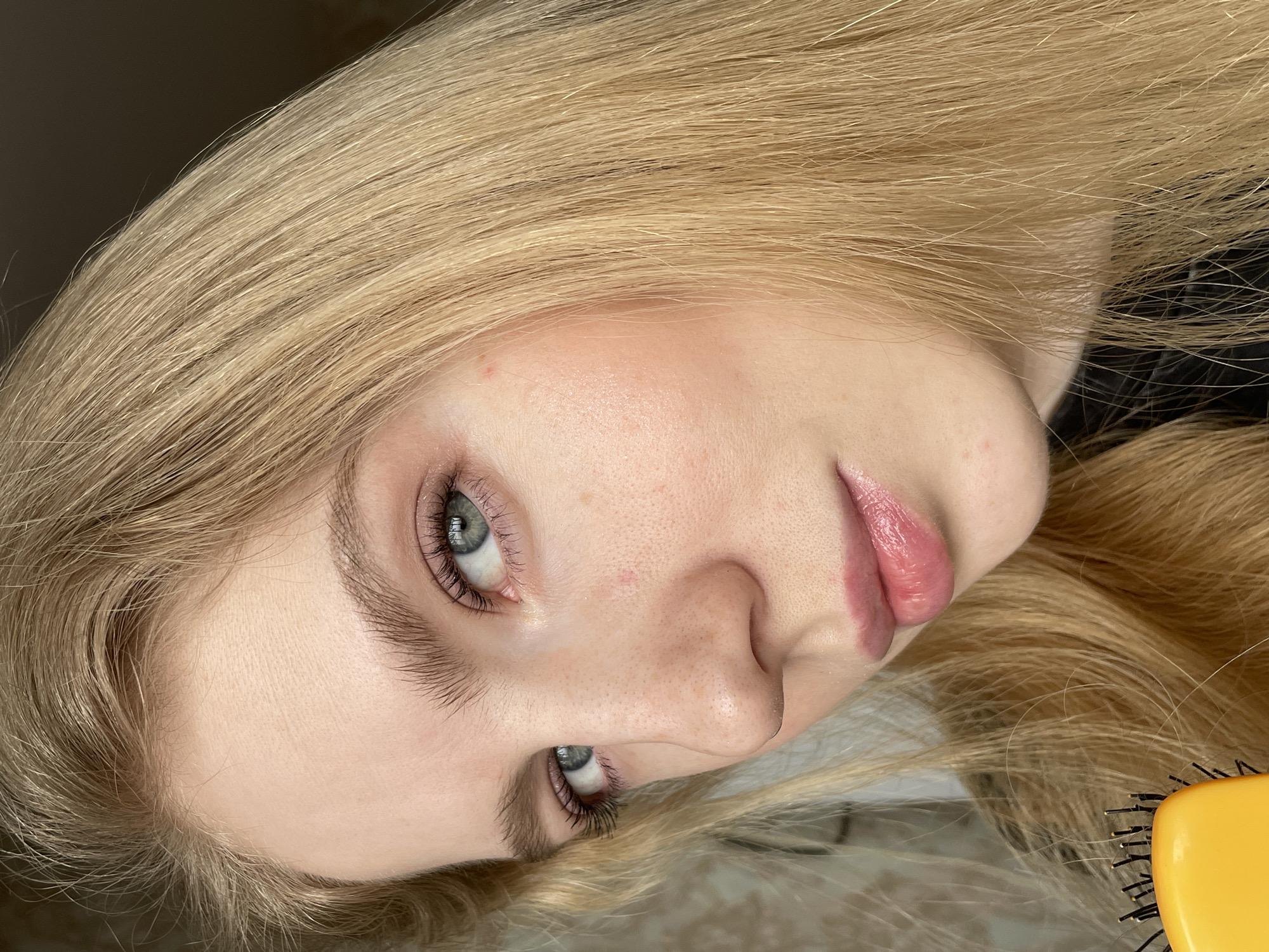 A close-up, tilted portrait captures a woman with long, blonde hair. Her skin is fair, and she has gray-blue eyes. Her lips are glossy. A yellow hairbrush is partially visible in the lower right corner. The location is not discernible from the image.