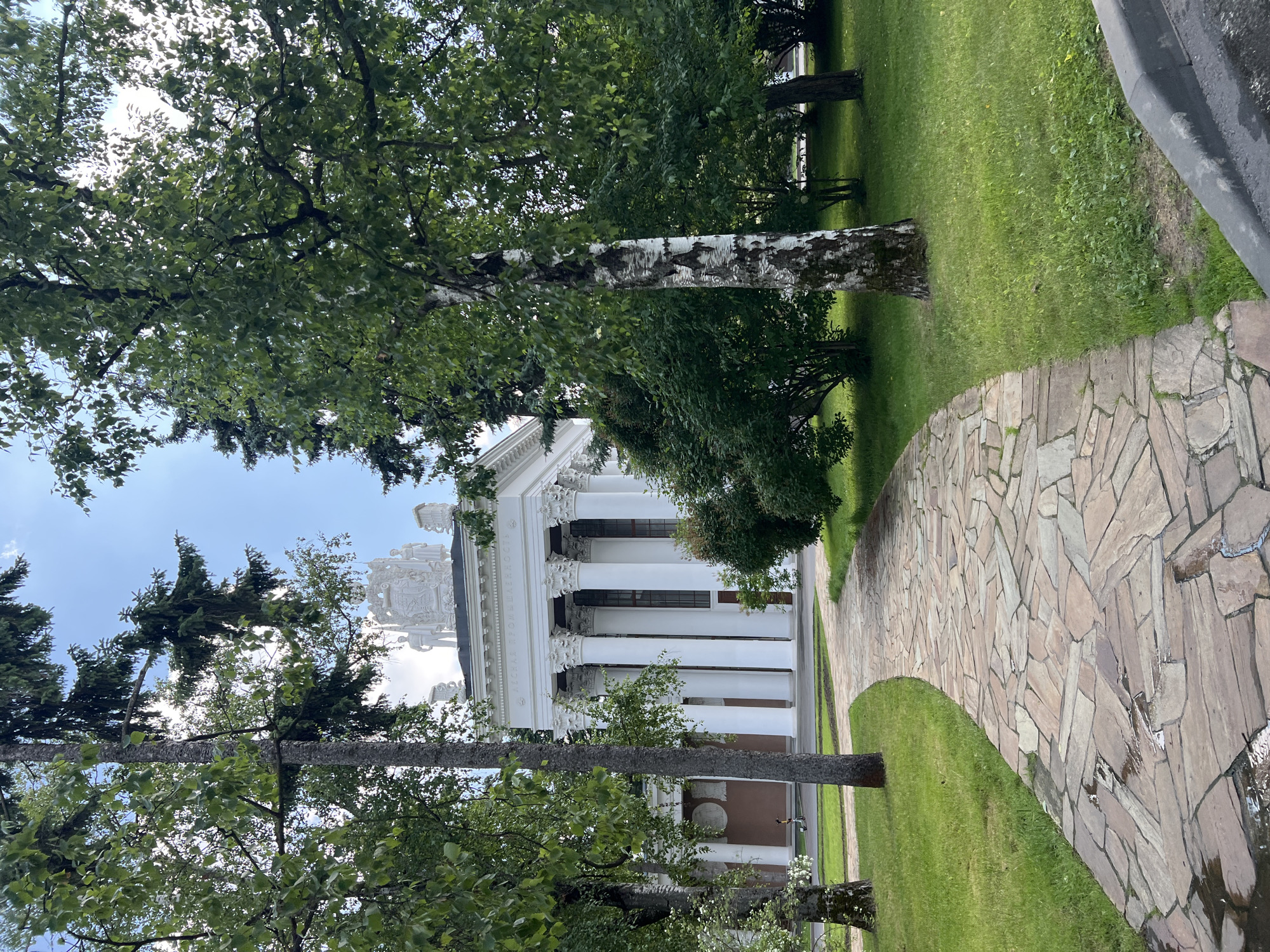 The image depicts a wide stone pathway leading towards a neoclassical building with a colonnade and ornate white sculptures on its roof. The pathway is lined with green grass and trees with lush foliage. A person is visible on a scooter to the left of the pathway, near the building's entrance. The architectural style and sculptures suggest that this location is likely within VDNKh (Exhibition of Achievements of National Economy) in Moscow, Russia, a park known for its Soviet-era pavilions and monumental art. The sky is partly cloudy, and the overall scene suggests a pleasant outdoor environment.