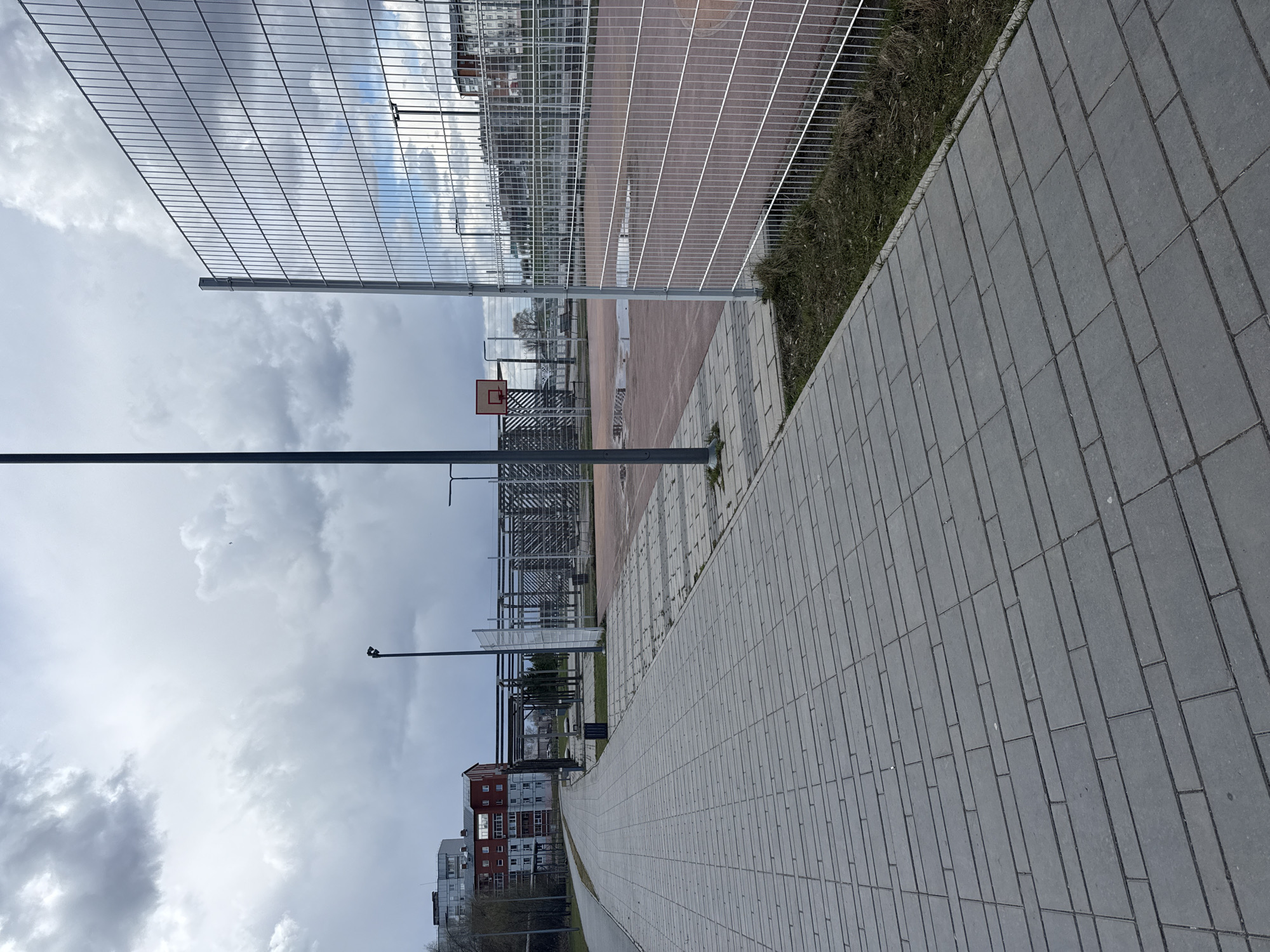 The image displays an outdoor urban recreational area in Votkinsk, Russia, under an overcast sky. A wide, grey paved pedestrian path, constructed from rectangular interlocking pavers, dominates the right side of the frame, extending into the distance.
To the left of the path, a narrow strip of dry grass and dirt separates it from a tall, metallic wire mesh fence. This fence encloses a sports court, likely a basketball court, given the presence of a basketball hoop with an orange rim and white backboard visible in the mid-ground. The court surface appears reddish-brown, and several reflective puddles on its surface suggest recent rain or wet conditions.
Beyond the sports court and fence, multi-story residential buildings of varying architectural styles, some with distinctive red roofs, line the distant horizon. Metal framework structures, possibly indicating another facility or an ongoing construction project, are also scattered in the background. Several tall, dark poles, likely lampposts or light fixtures, are positioned throughout the scene.
The sky is heavily overcast with dense, grey clouds, indicating a cloudy daytime setting with no direct sunlight. The overall atmosphere is quiet and still, as no people or activity are visible in the scene. There is no discernible text present in the image.