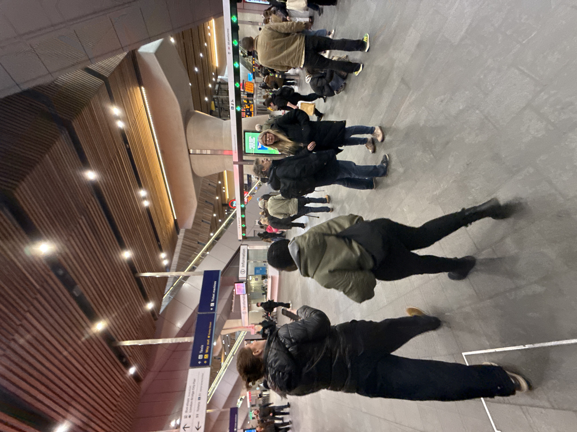 The image depicts a bustling scene inside a transportation hub, likely a railway station, in London, United Kingdom. A multitude of people are seen walking, indicating a high traffic flow. The location appears to be London Bridge, indicated by the sign "London Bridge City Pier," which suggests it is a train station. Signage for "Taxis," "Way out," "Ticket machines," and "All platforms" further support the location being a transportation hub. The interior architecture includes a distinctive wooden ceiling, with rows of lights accentuating the design. People are dressed in casual, everyday clothing, indicating a normal day and time rather than a special event. The floor is light gray. The overall impression is of a crowded, busy transport hub, with people going about their travel routines.