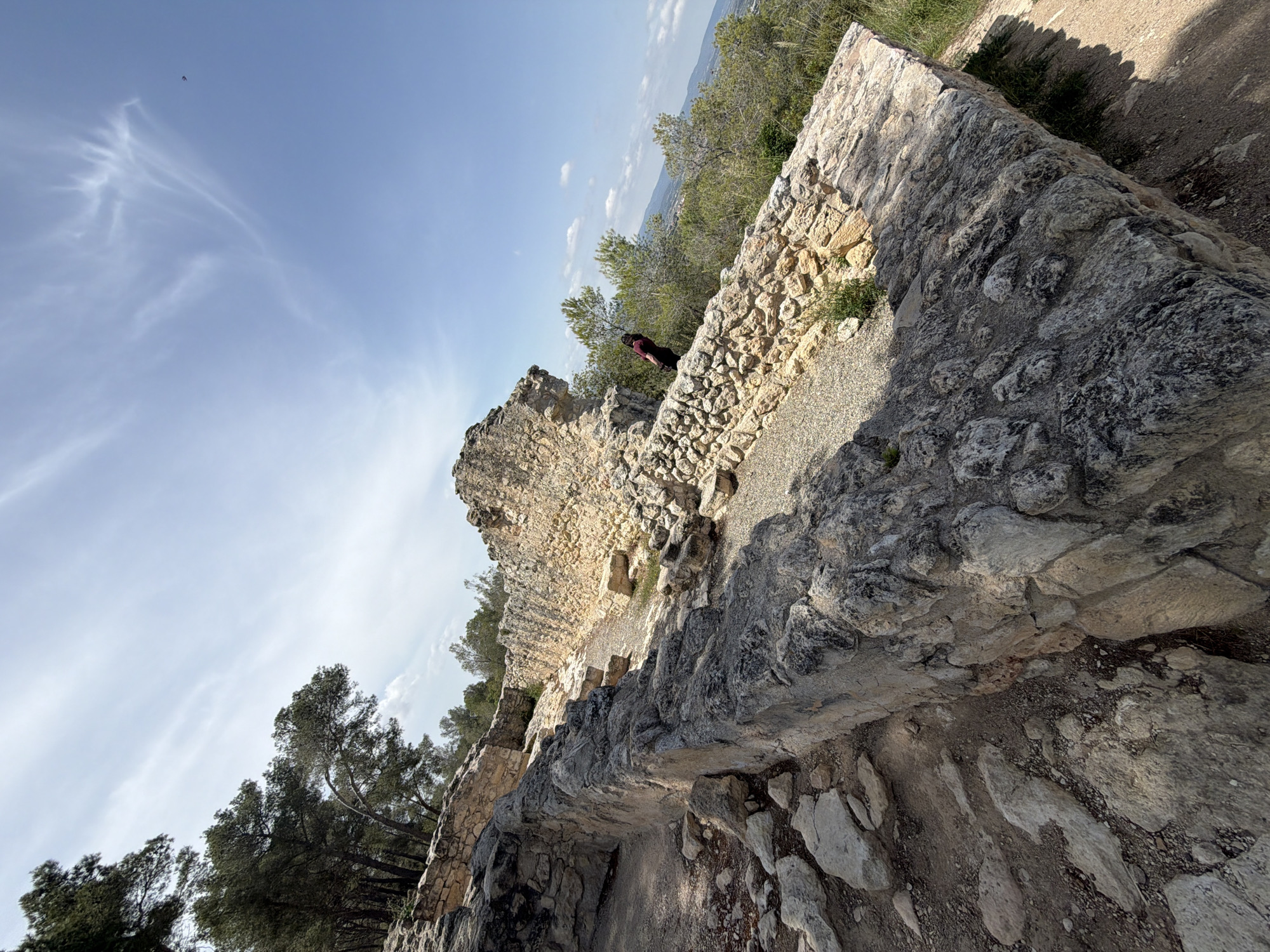 The image presents an outdoor scene depicting ancient stone ruins or walls under a bright, partly cloudy sky. In the immediate foreground, dark, textured stone blocks form a rugged wall or structure, with smaller stones and dirt on the ground around it. Moving into the midground, a substantial, lighter-colored stone wall meanders, forming part of what appears to be a historical fortification or building. A gravel path runs behind this wall.
A person wearing a dark top is partially visible behind the midground stone wall, looking out over the landscape. Their upper body and head are visible against the backdrop of distant greenery. Beyond the stone structures and the person, numerous evergreen trees, possibly pines, dot the hilly terrain. In the far background, a vast, hazy landscape of rolling hills or mountains stretches towards the horizon under a pale blue sky.
The sky is largely clear, with some delicate, wispy white clouds trailing across it. A small, dark, indistinct object, possibly a bird or a distant aircraft, is visible high in the sky. The strong sunlight casts sharp shadows across the stone surfaces, indicating a sunny day, likely late morning or early afternoon. The overall impression is one of exploring an ancient site with panoramic views of the surrounding natural environment, consistent with a historical location like Olèrdola, Spain. There is no visible text in the image.