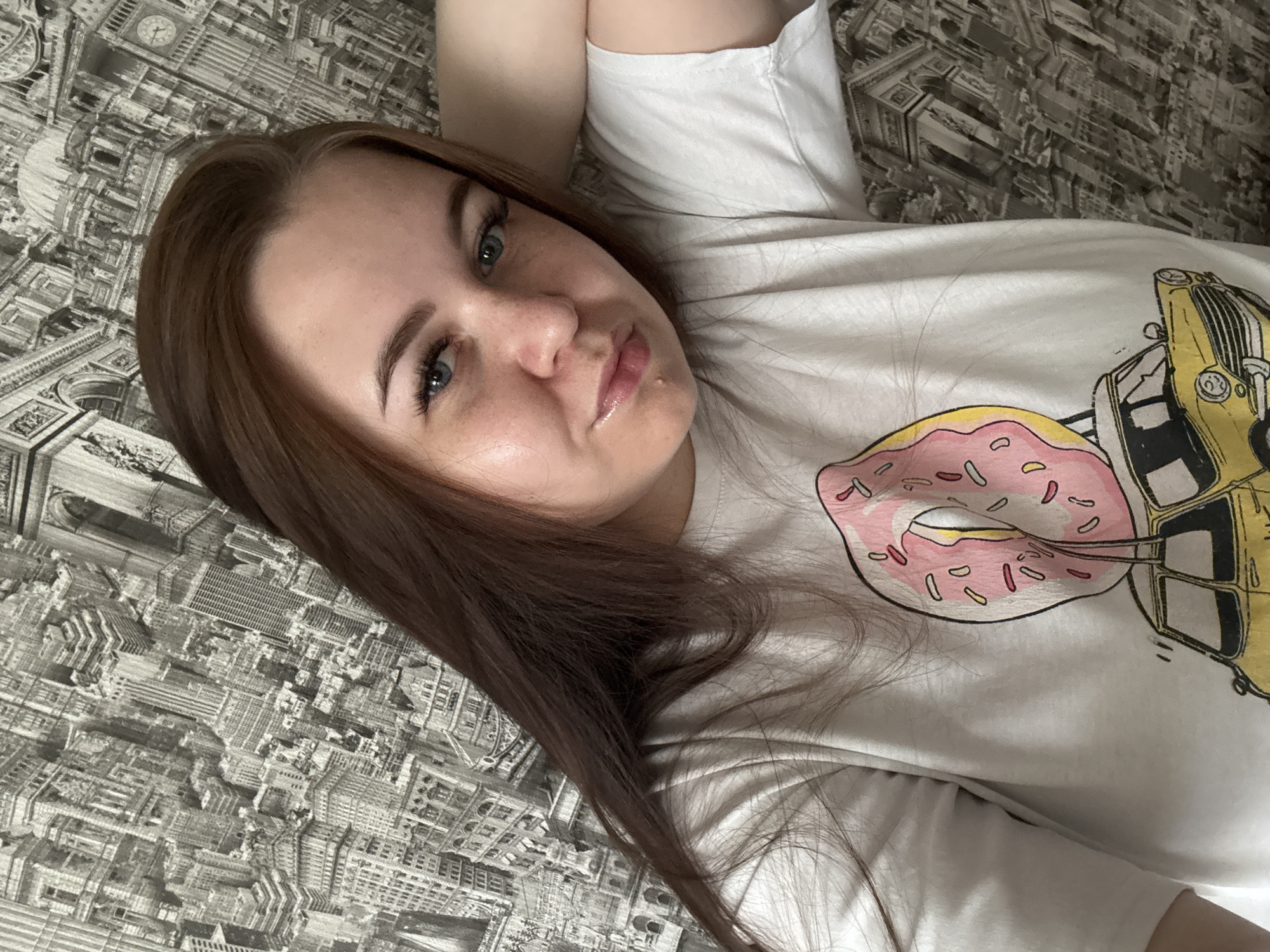 A woman with long, brown hair lies on a bed with a gray cityscape-patterned comforter. She is wearing a white T-shirt with a design featuring a pink donut and a yellow taxi. The scene takes place indoors, but there are no immediate indications of it being Chaikovsky, Russia.