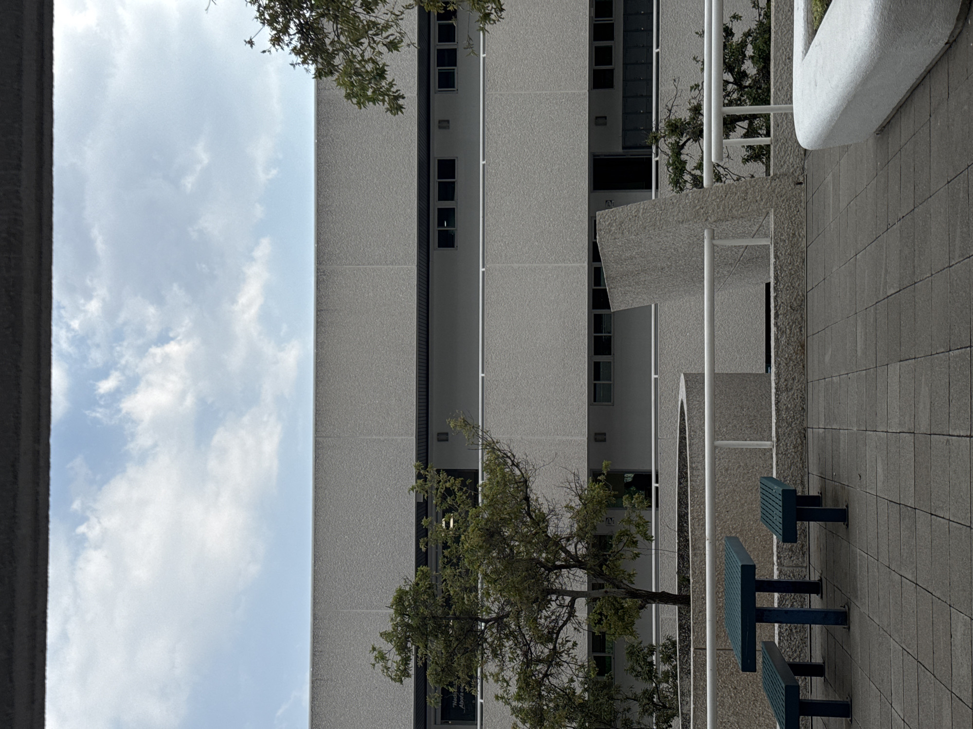 This image captures an outdoor daytime scene within an urban or campus setting in Puebla, Mexico, viewed as if looking upwards from a lower vantage point, with the image rotated 90 degrees counter-clockwise. The central subject is a tall, light-grey building with a textured facade, featuring numerous vertical rows of rectangular windows, some appearing dark and reflective. A darker vertical strip with additional windows runs along one section of the building.
In the foreground and midground, a paved area with grey square tiles is visible, furnished with several dark blue or green picnic tables and benches, suggesting a public seating area. White vertical posts, likely part of railings or structural supports for overhangs, are prominent. A large, light-grey concrete structure with a rounded, organic shape, possibly a planter or decorative architectural element, is situated towards the right.
Throughout the scene, several trees with green foliage provide natural elements, with branches extending near and in front of the buildings. Through the gaps between the structures, a bright sky with scattered white clouds is visible, indicating clear daytime weather. The scene is devoid of people or explicit activity, presenting a calm, architectural landscape. Faint, partially obscured white text, possibly including the word "Abierta" (Spanish for "open"), is barely discernible on a dark panel of the building, partially hidden by tree leaves.