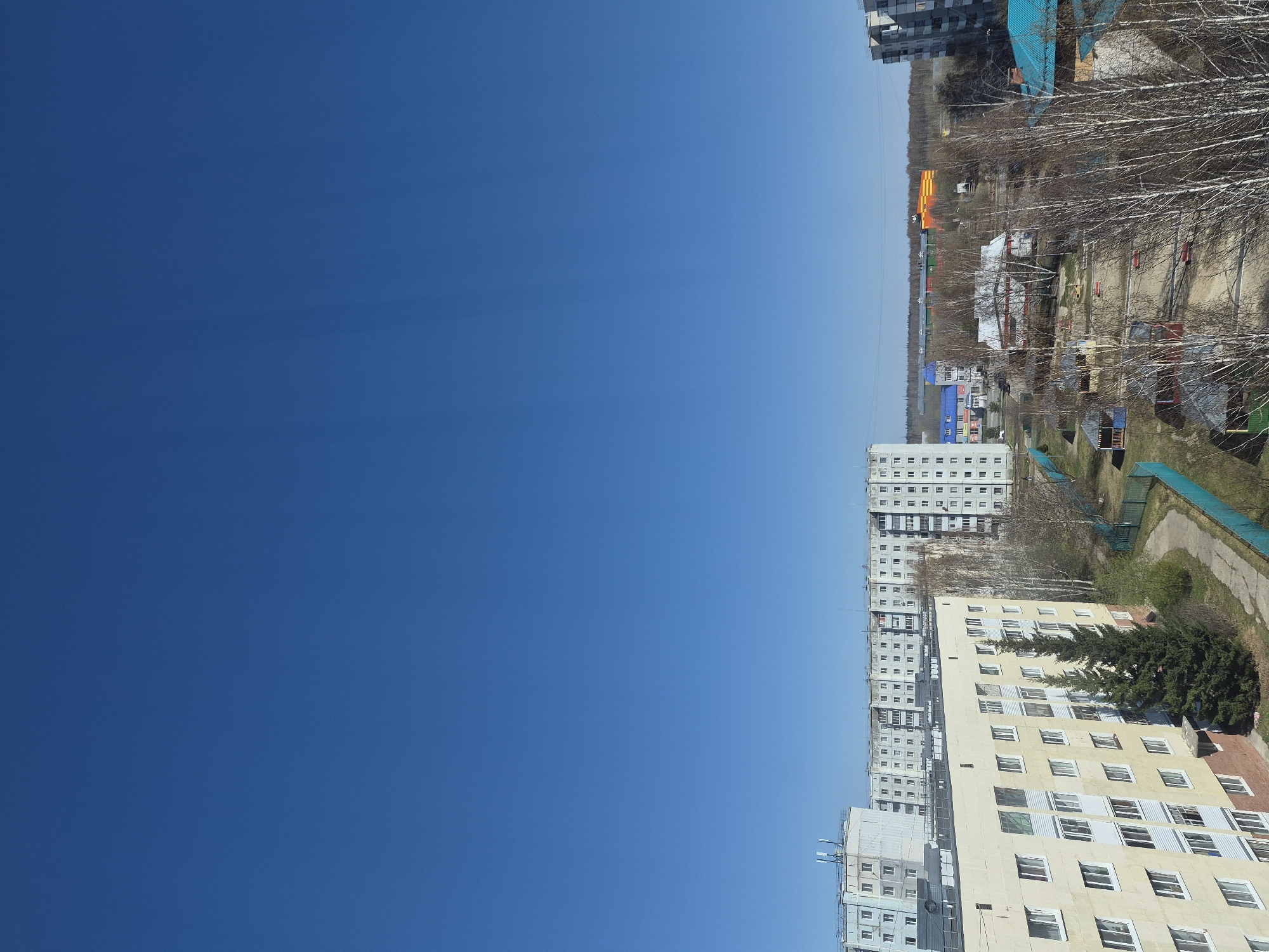 The image shows a high-angle view of a cityscape in Tobolsk, Russia, under a clear blue sky. Buildings of various heights, including apartment blocks, are visible, as well as playground and park areas, alongside bare trees.